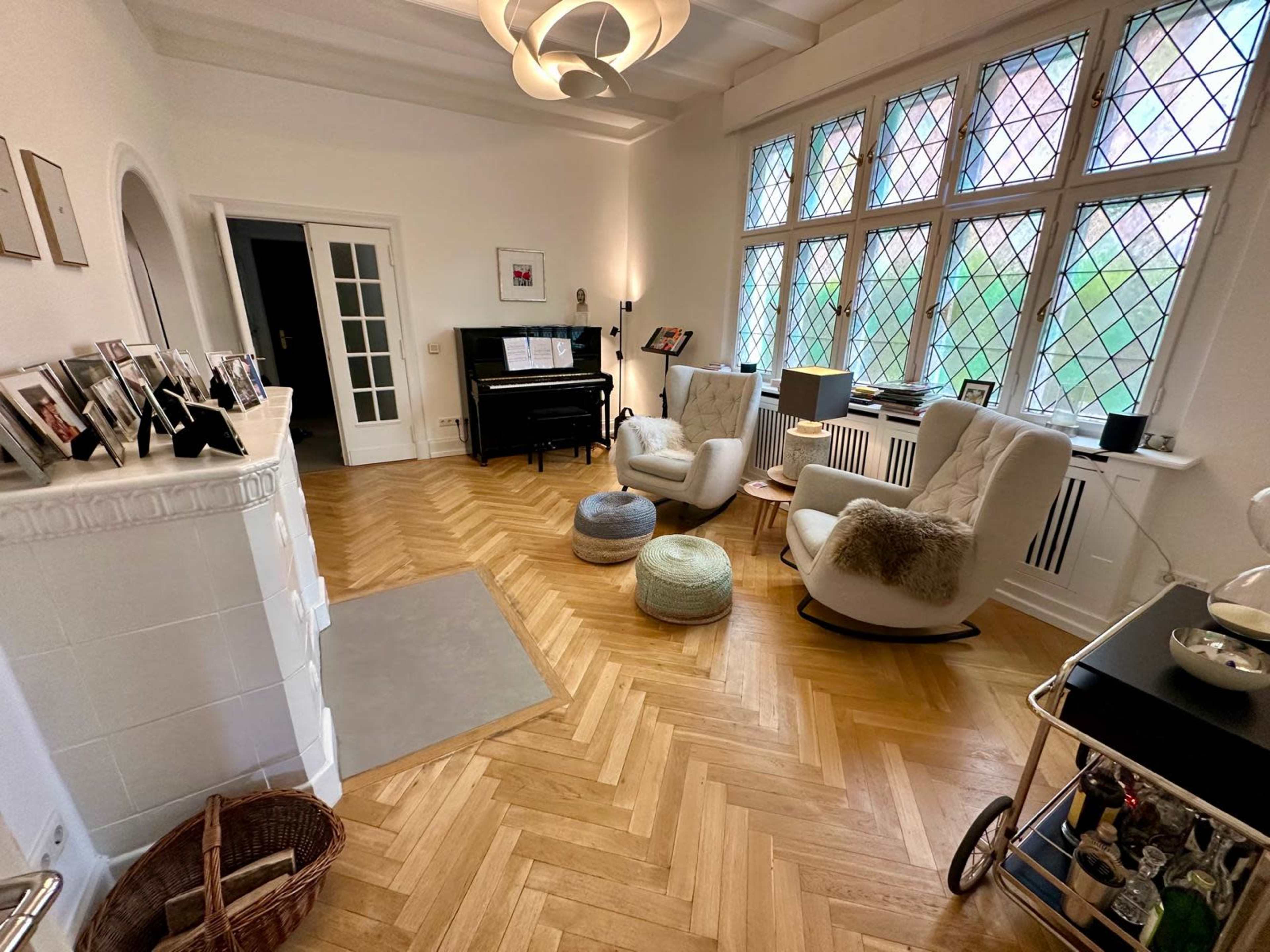 The image shows a spacious living room with two armchairs, a piano, and large windows featuring diamond-shaped panes.