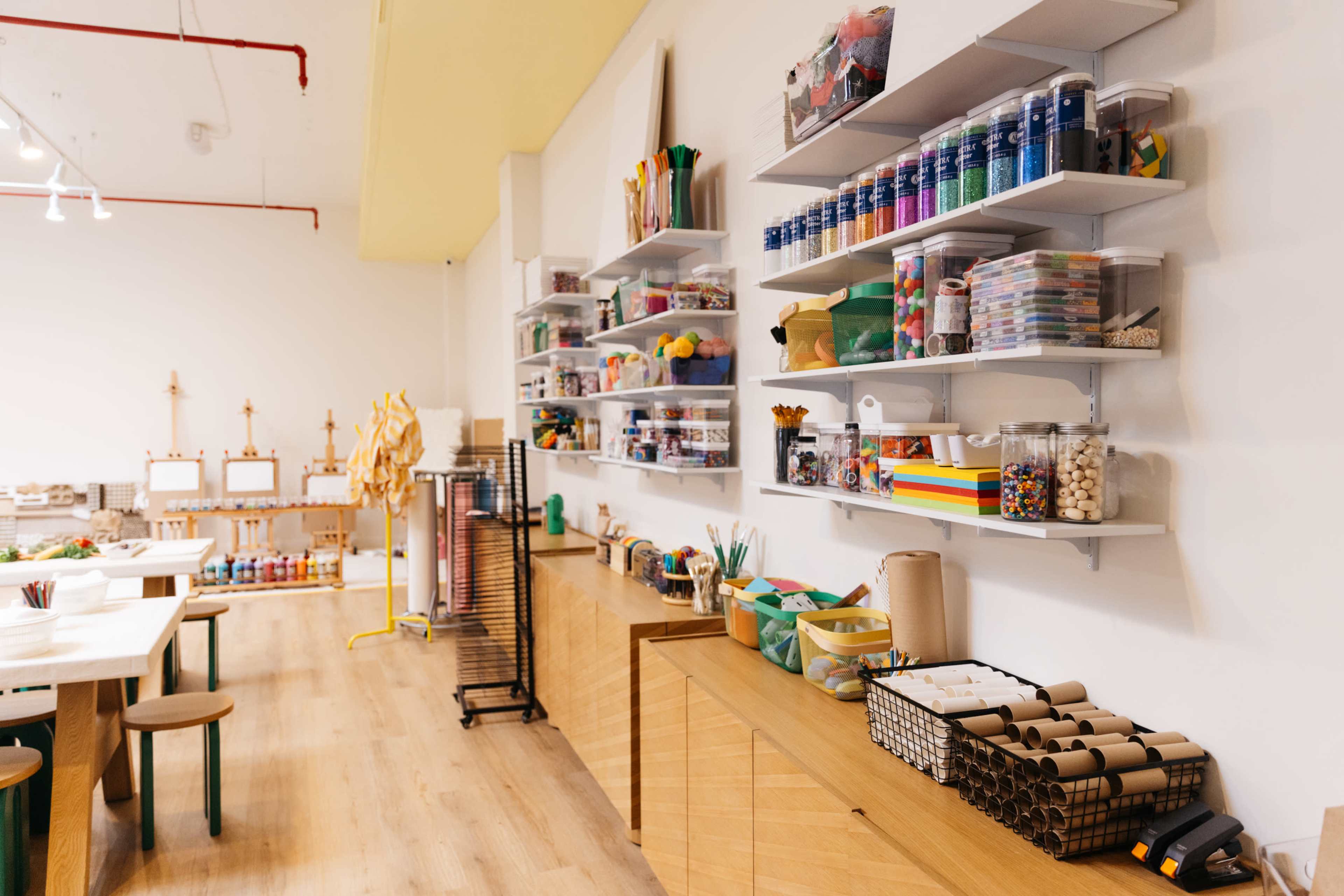 The image shows a well-organized craft room with shelves stocked with art supplies, including paints, paper, and various crafting materials, alongside wooden tables for creative activities.