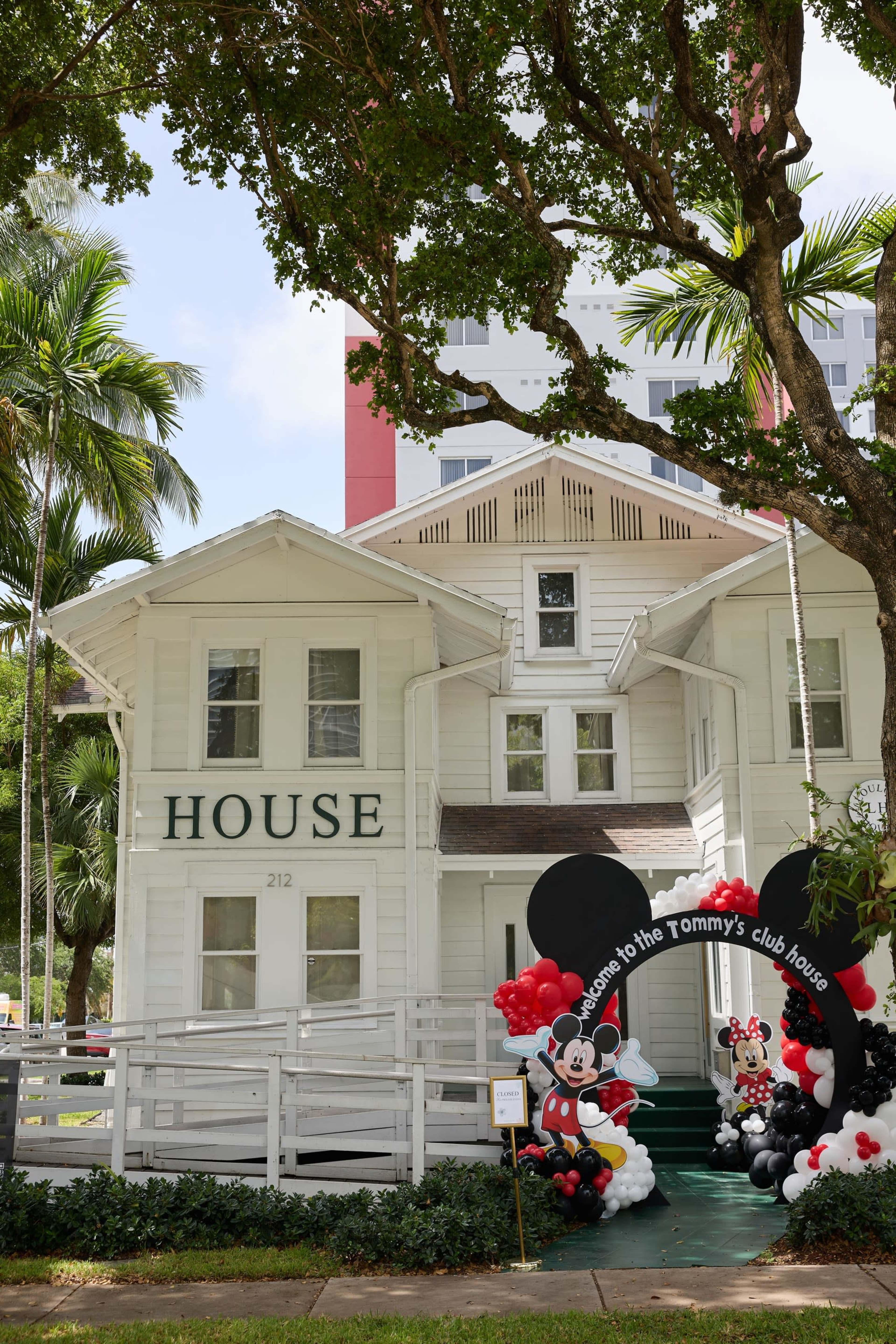 The image shows a white, two-story house with the word "HOUSE" prominently displayed on its front, accompanied by a decorative arch featuring Mickey and Minnie Mouse-themed balloons.