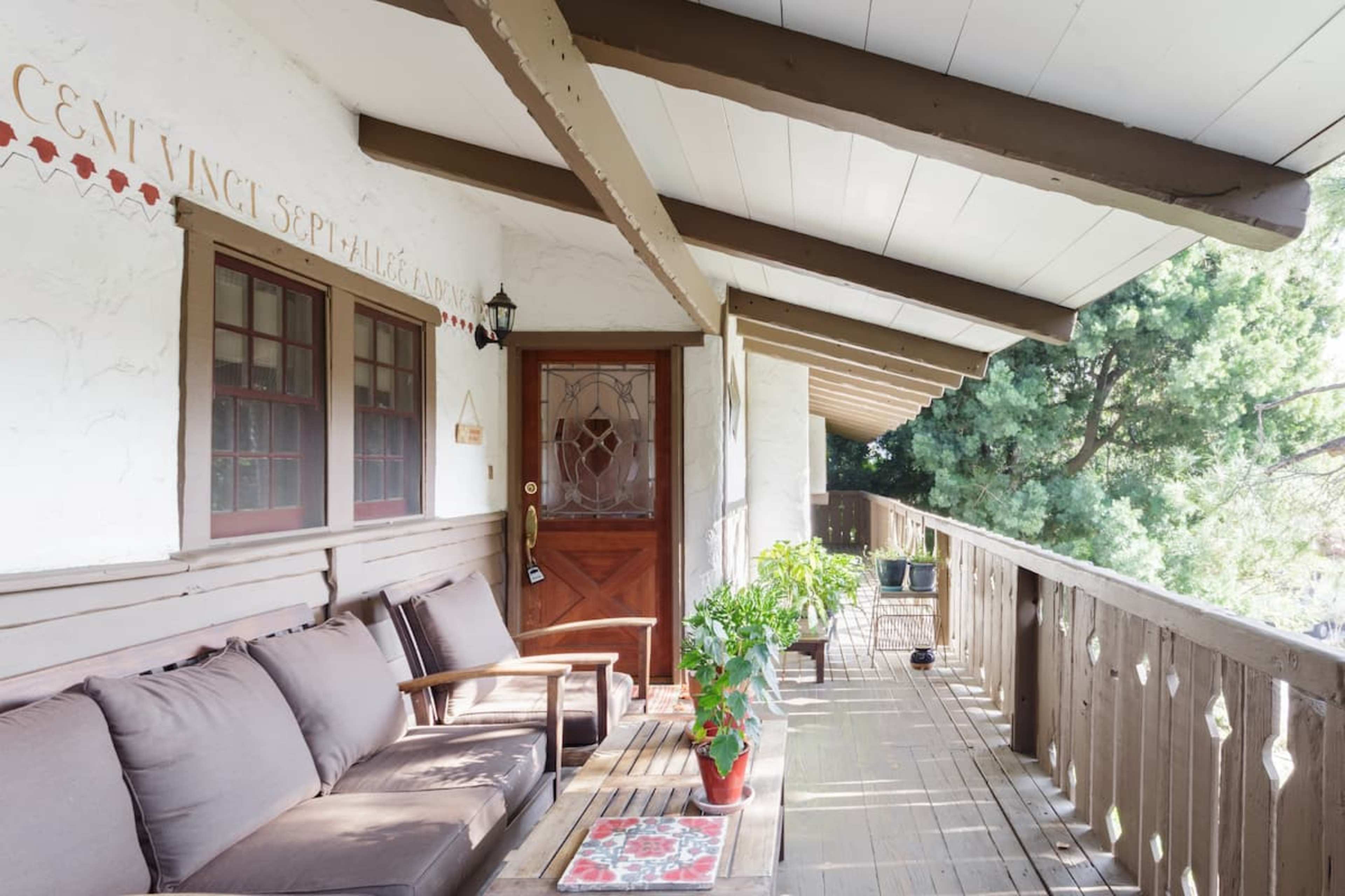 A wooden porch with a set of brown cushions, a potted plant, and a door at one end, surrounded by trees.