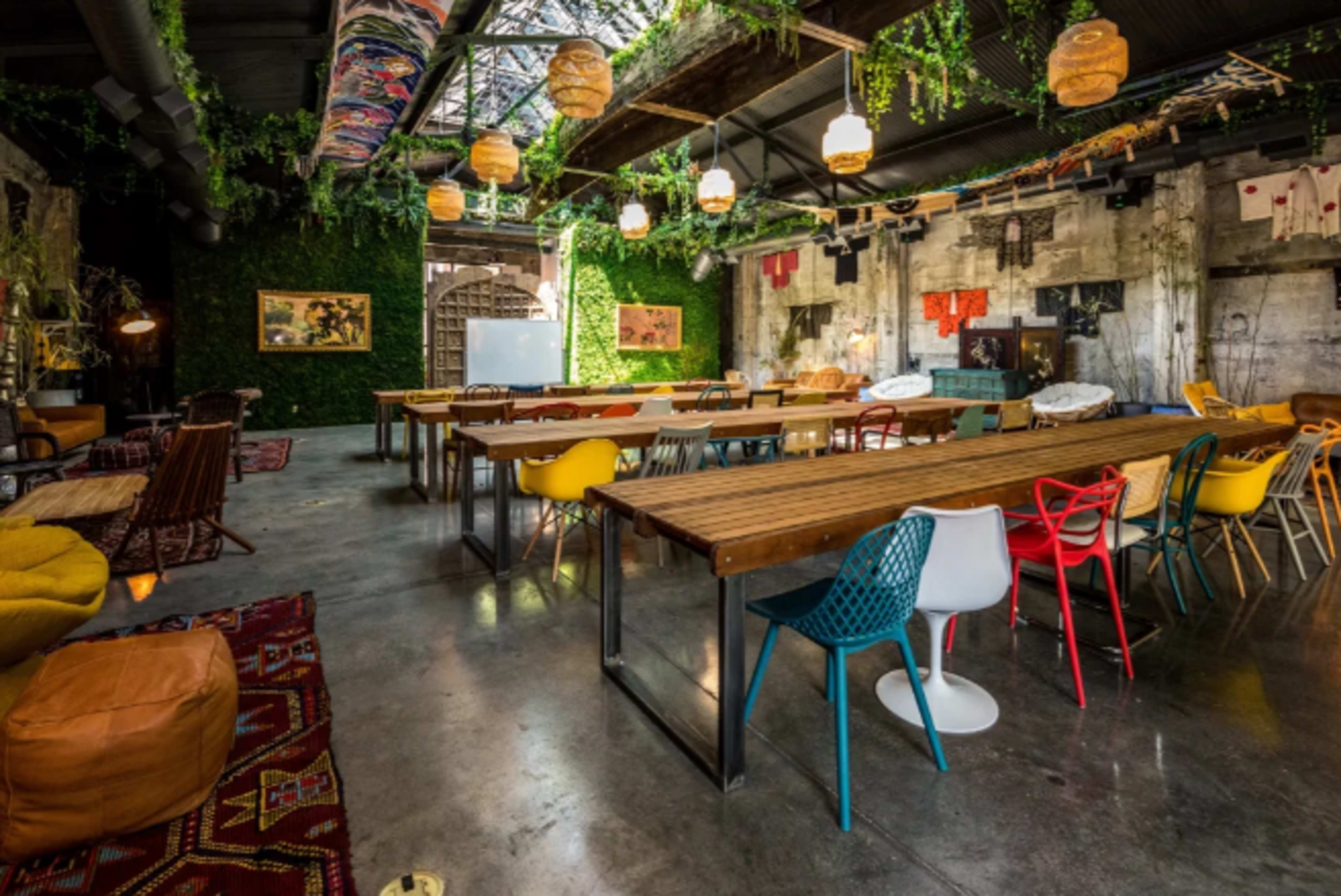 The image shows a spacious indoor venue with long wooden tables surrounded by colorful chairs, decorative plants, and eclectic artwork on the walls.