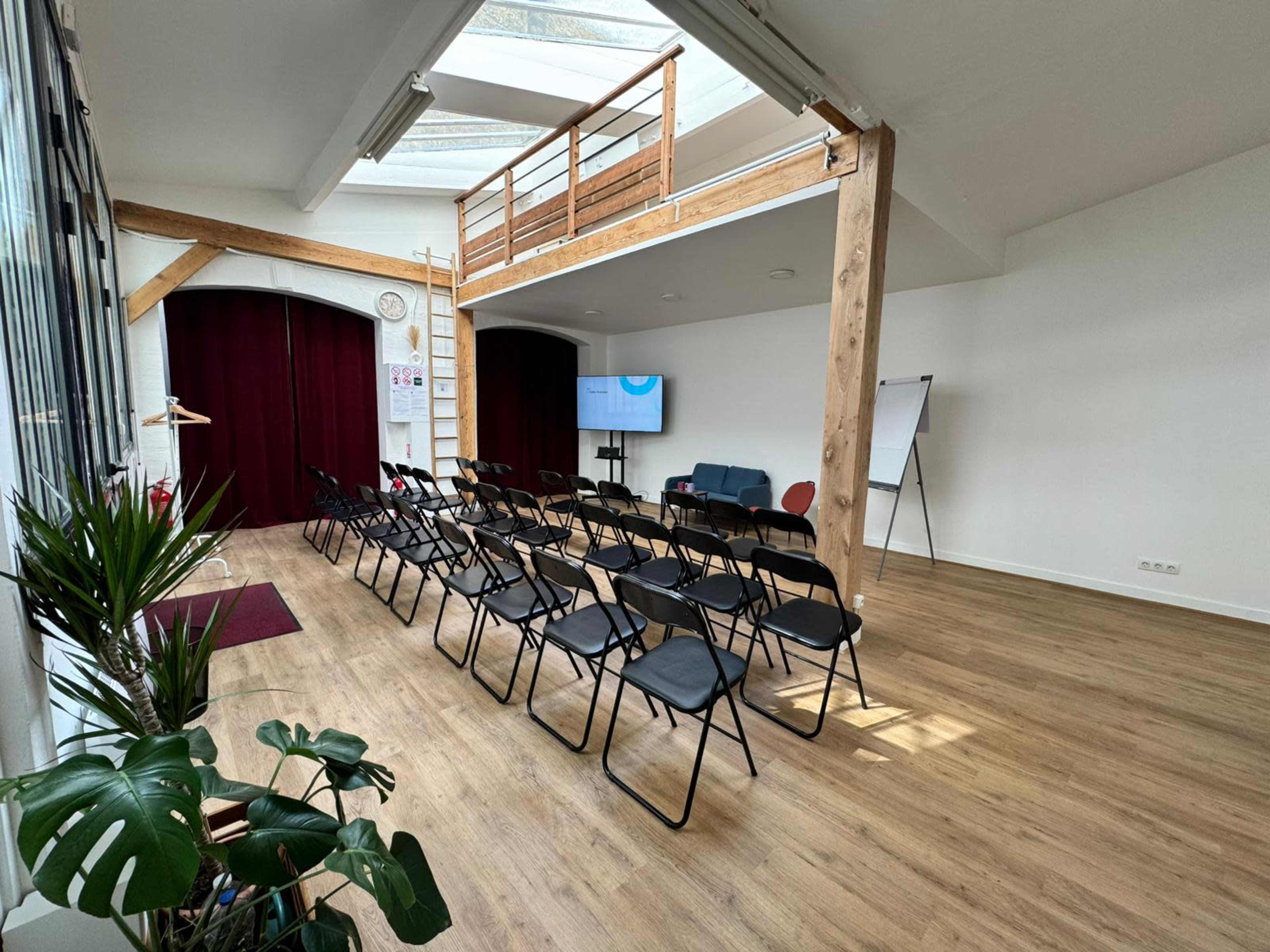 The image shows a spacious room set up for a presentation, featuring rows of black chairs facing a screen, with wooden beams and large windows letting in natural light.