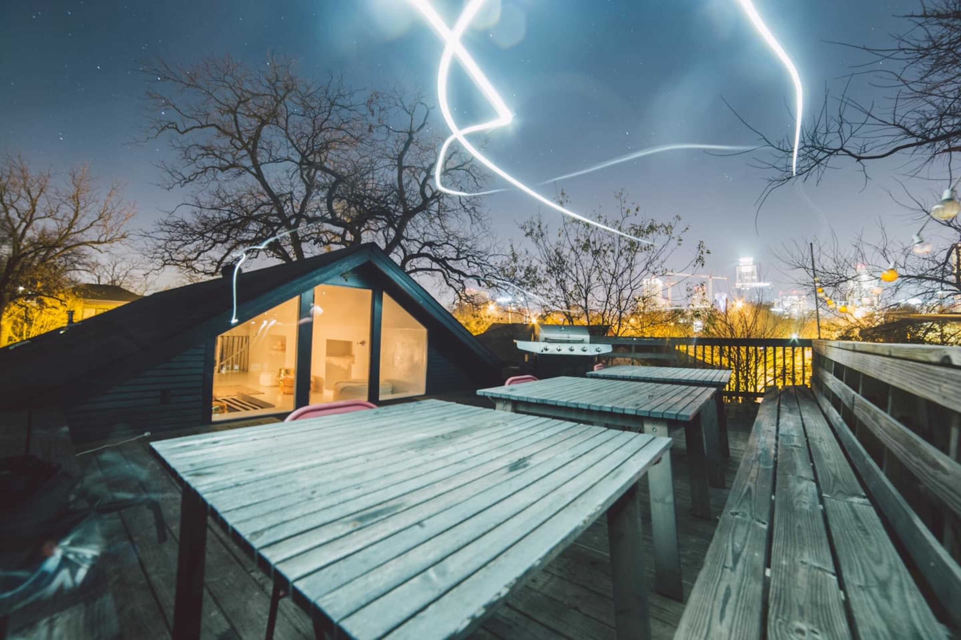 A rooftop deck with wooden tables and chairs, overlooking a city skyline illuminated at night, with light trails in the sky.