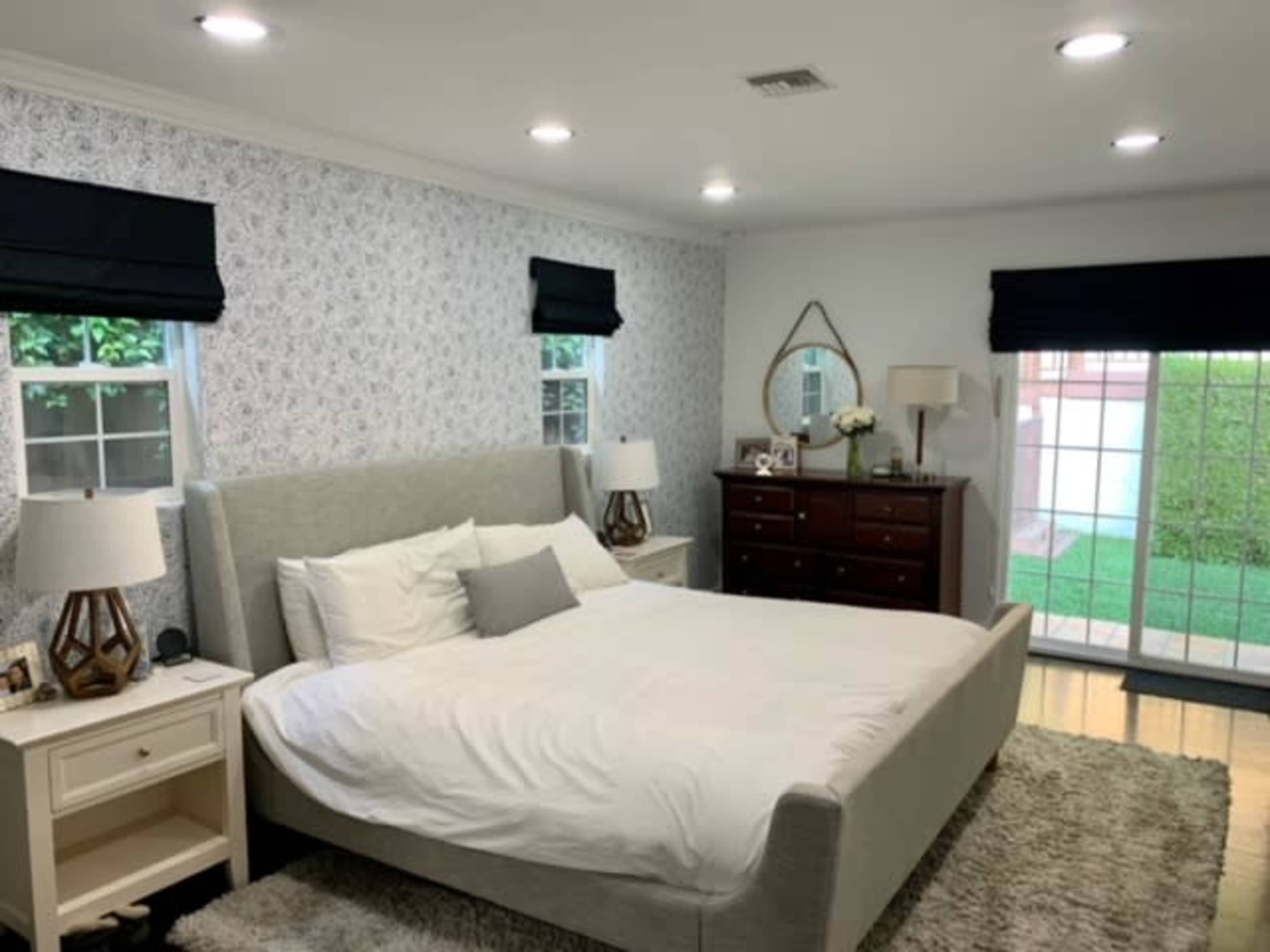 A spacious bedroom features a light-colored bed with two nightstands, a dresser, and large windows with black out curtains.