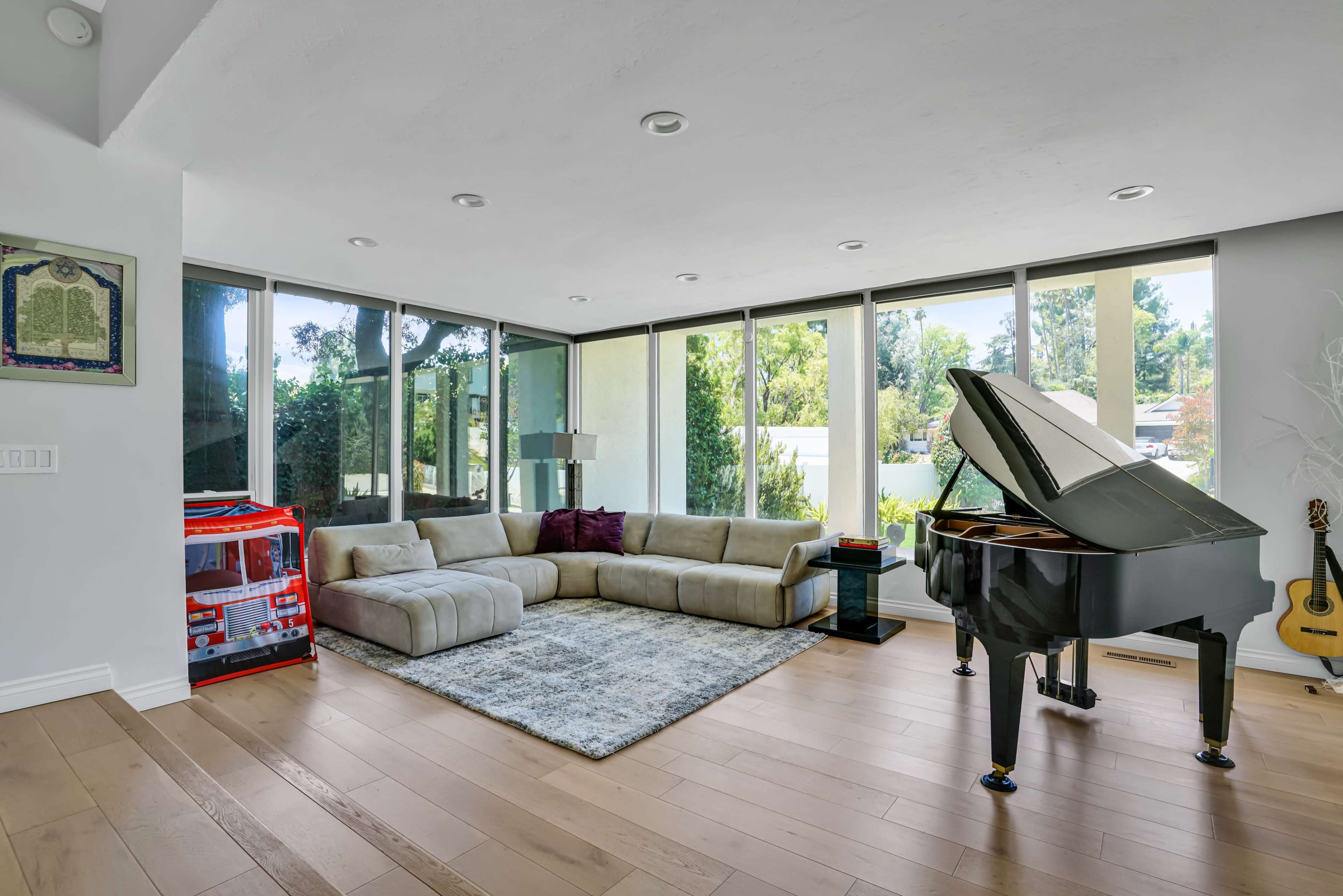 The image shows a modern living room featuring a large sectional sofa, a grand piano, and a toy storage unit near large windows that provide outdoor views.