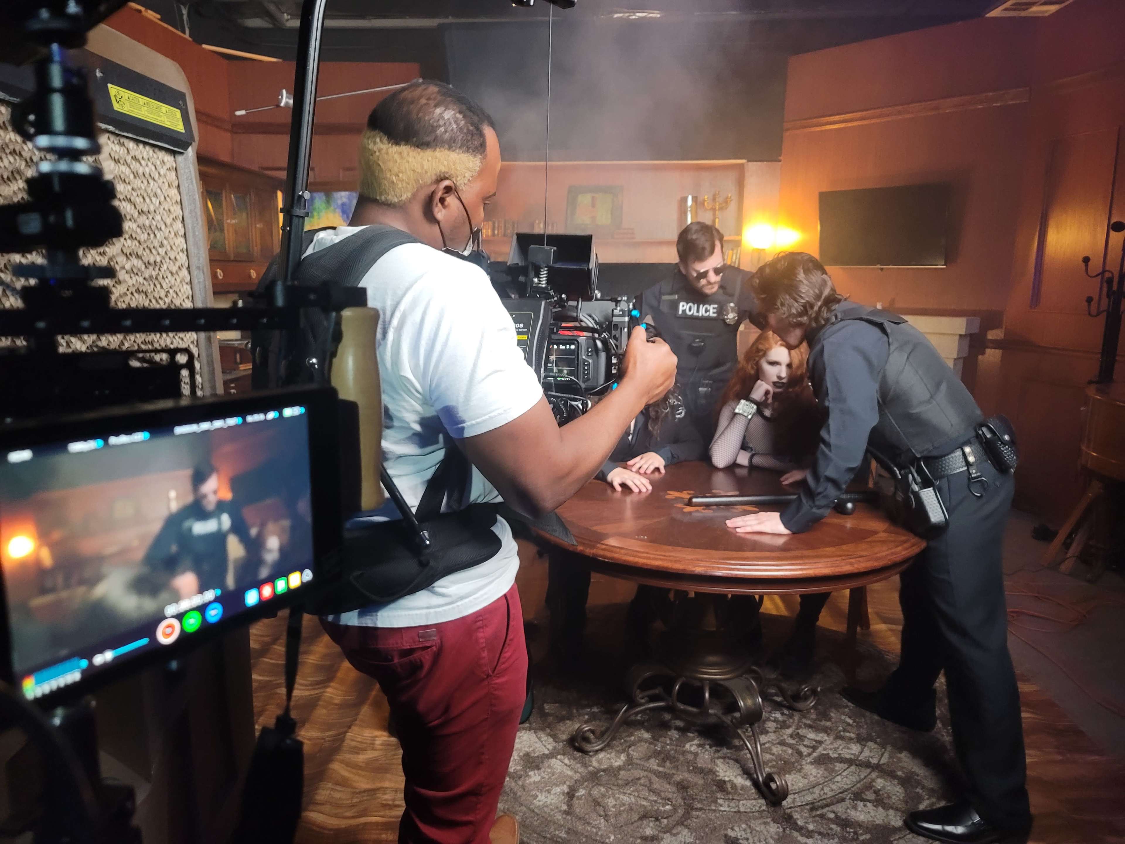 A film crew captures a scene with police officers and a suspect gathered around a table in a dimly lit room.