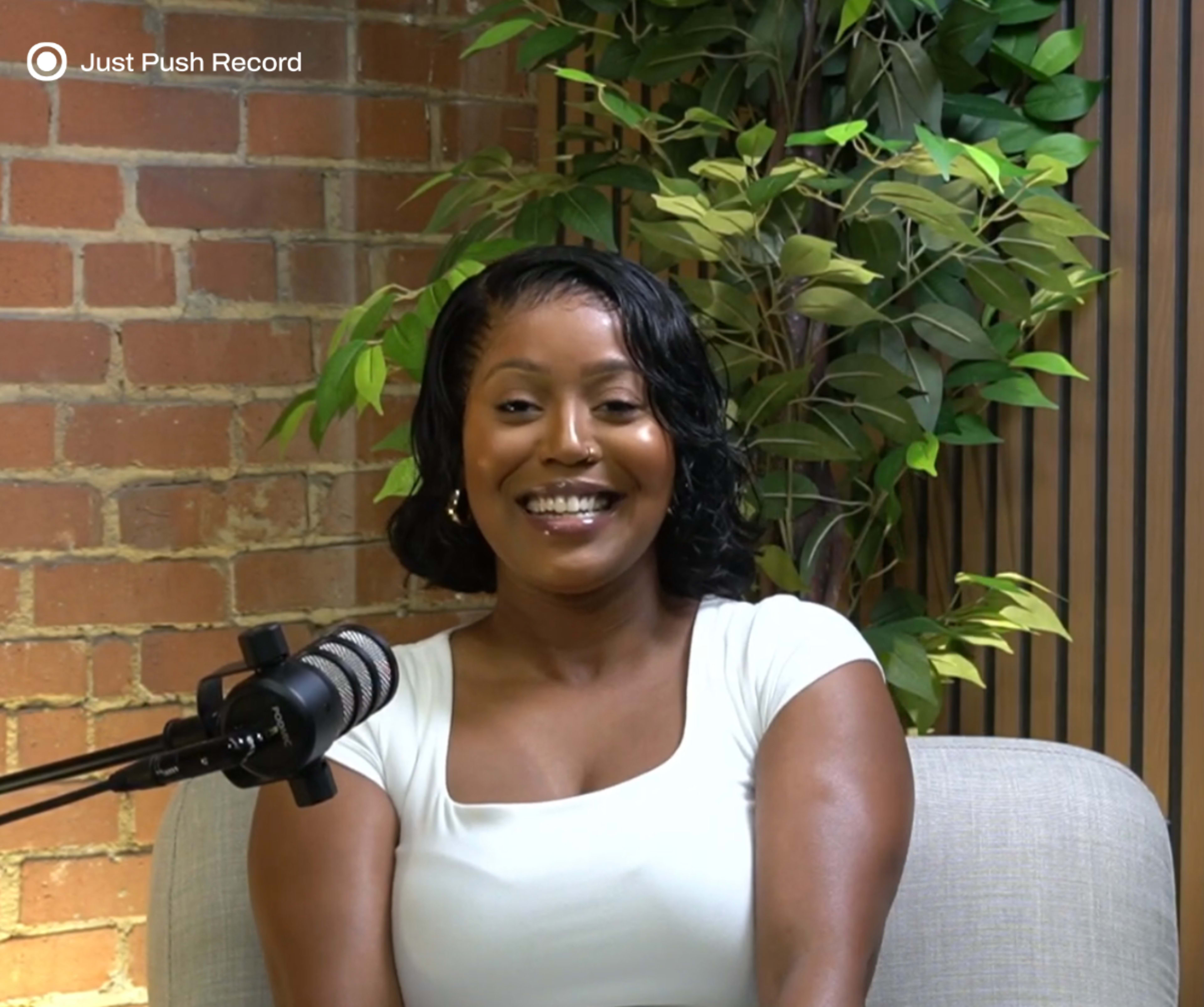 A woman is sitting on a couch in front of a microphone, with a brick wall and greenery in the background.
