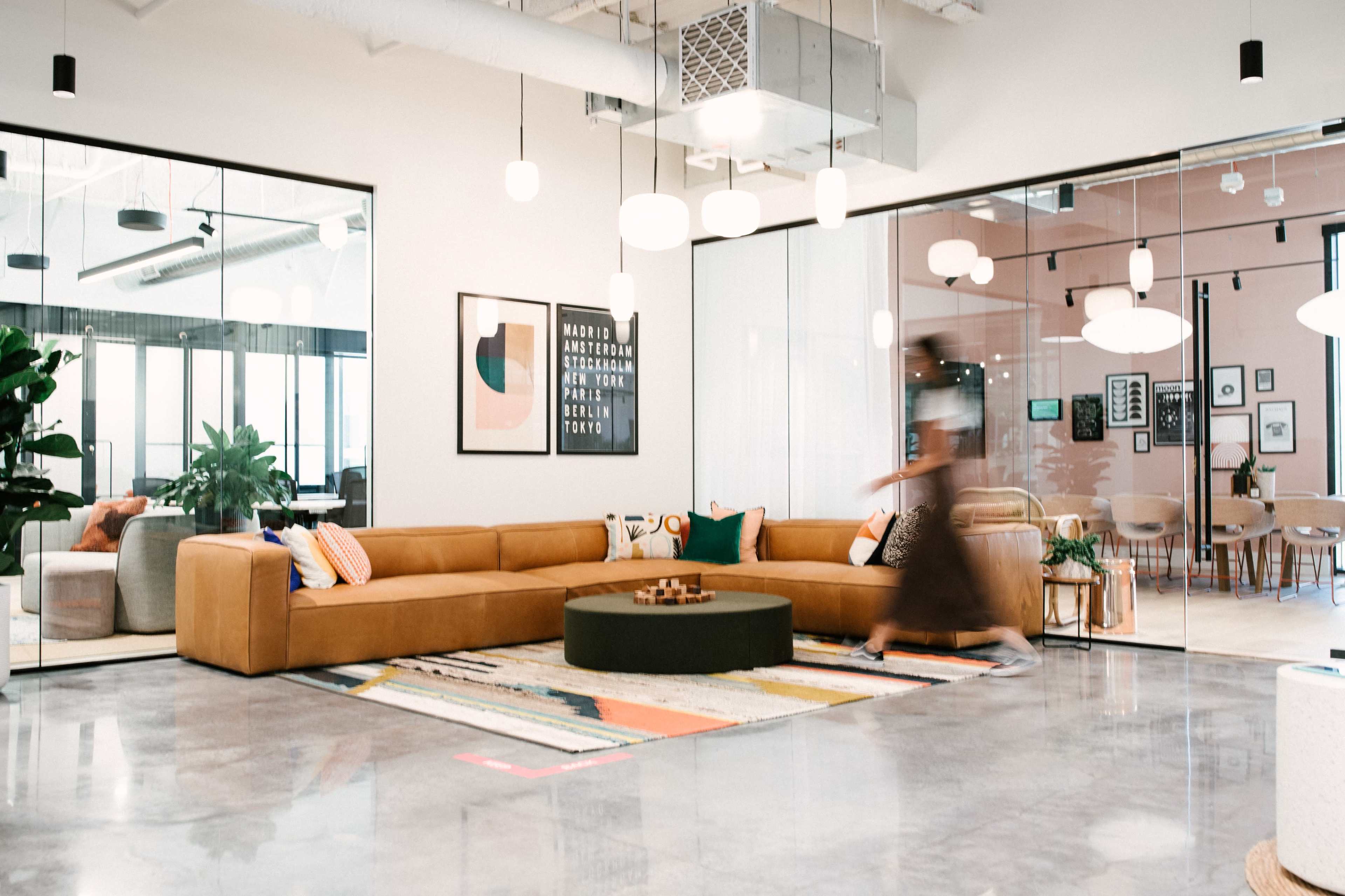 The image shows a modern office lounge area with a large brown sectional sofa, a circular coffee table, decorative plants, and colorful artwork on the walls.
