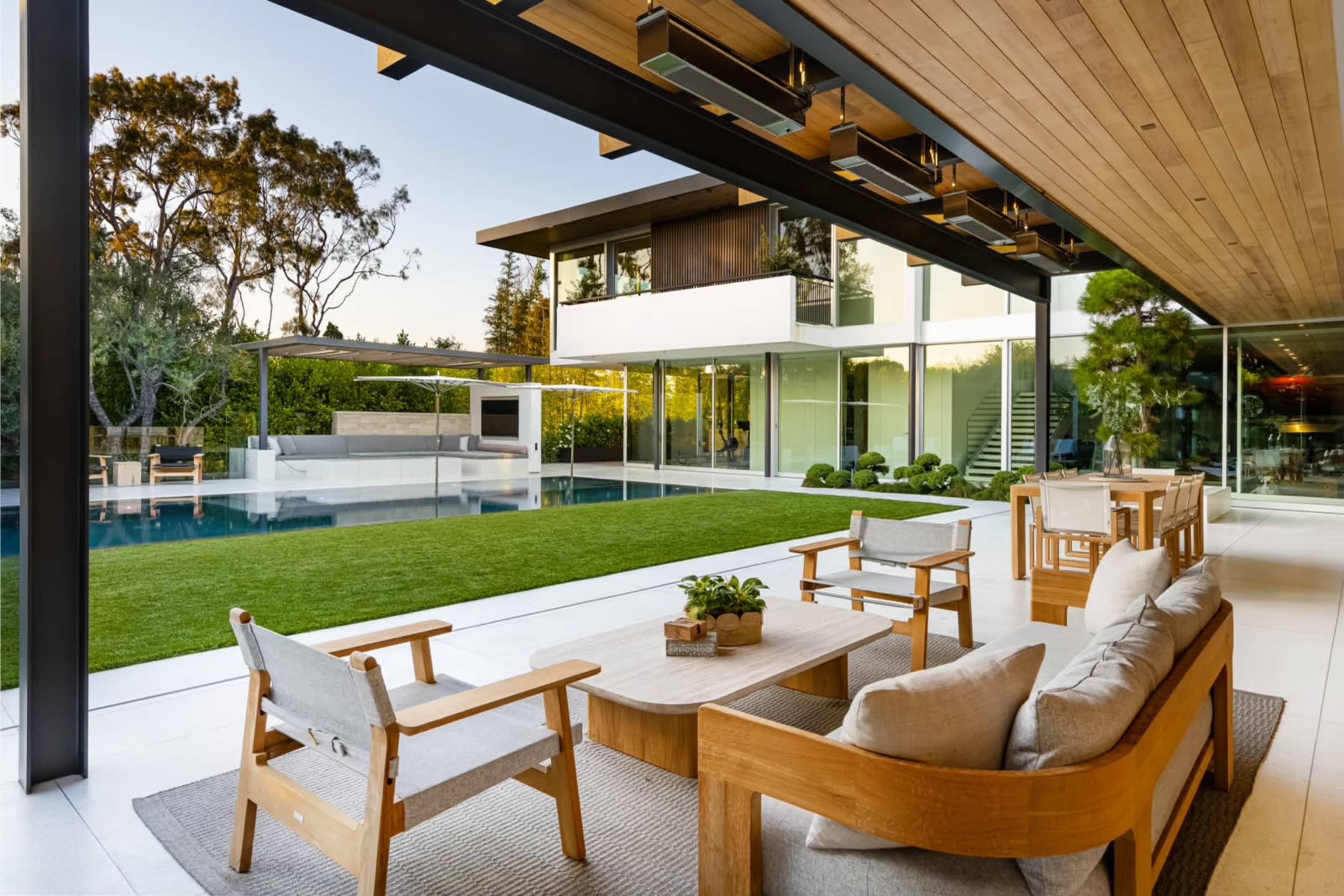 A modern outdoor living space features a sleek seating arrangement, a wooden table, and a view of a swimming pool surrounded by landscaped greenery.
