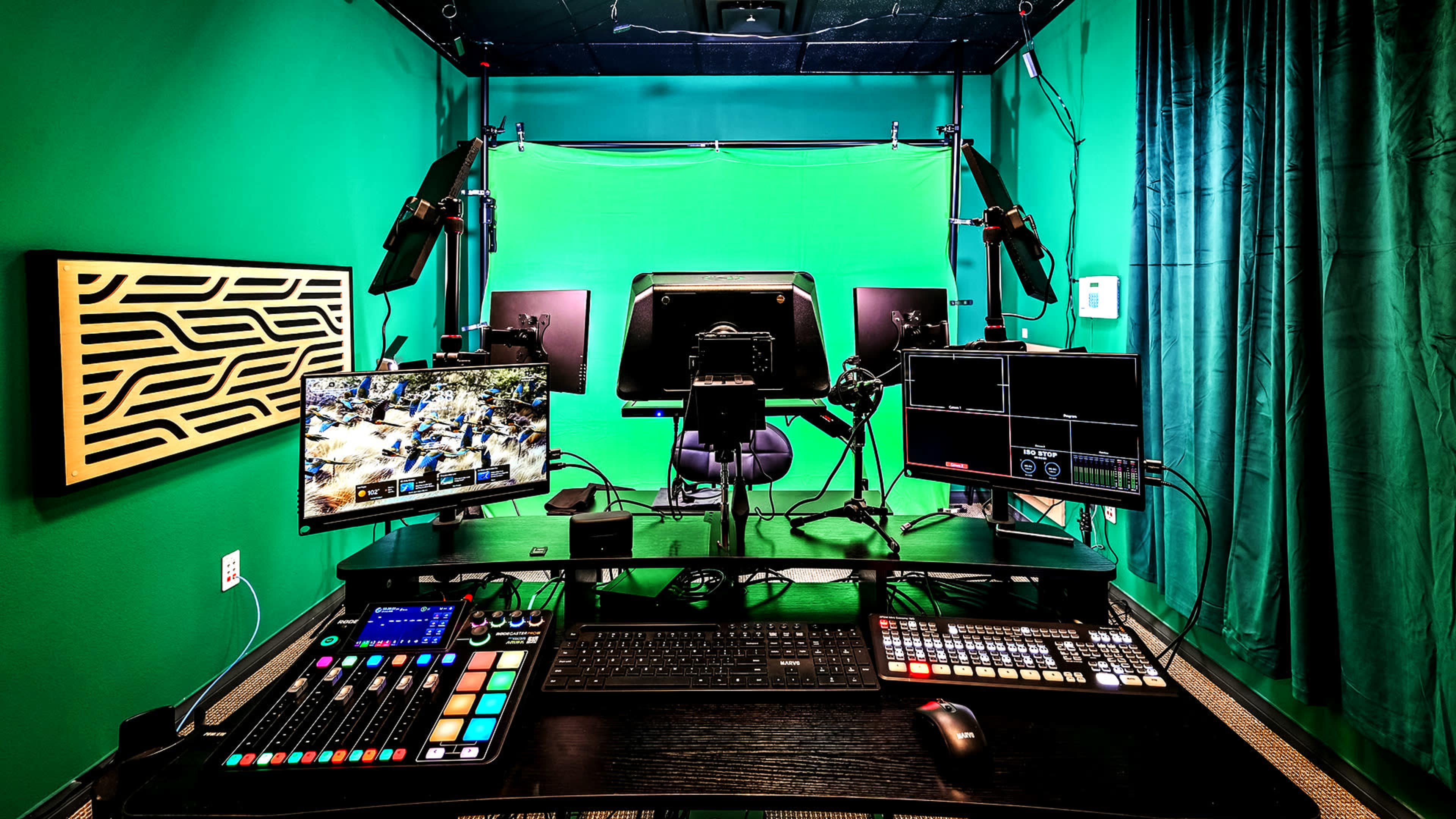 The image shows a professional video production studio with multiple monitors, control panels, and a green screen backdrop in a centrally placed work area.