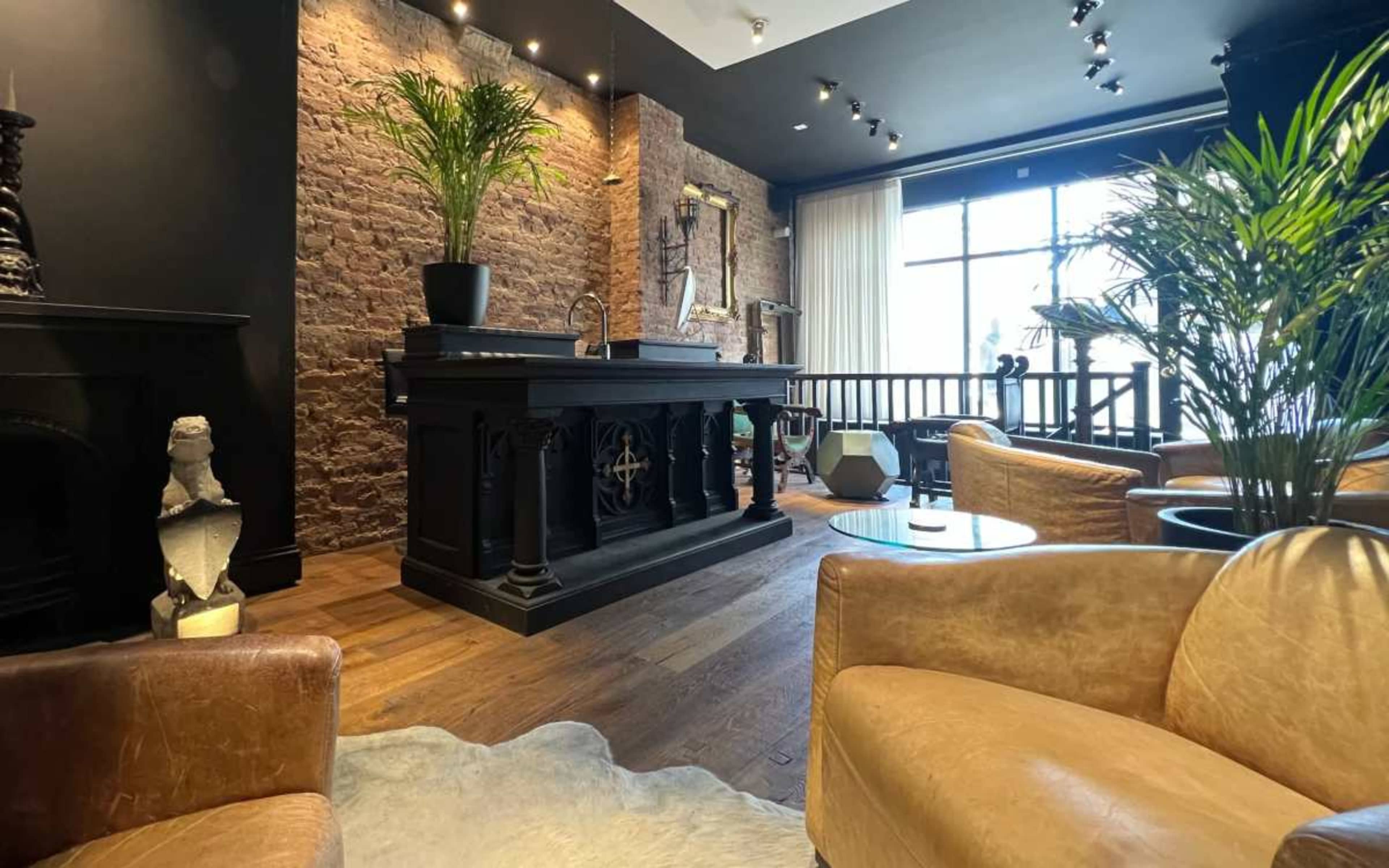 The image shows a stylish lounge area with a dark wooden bar, brick walls, and leather seating arranged around a small table.