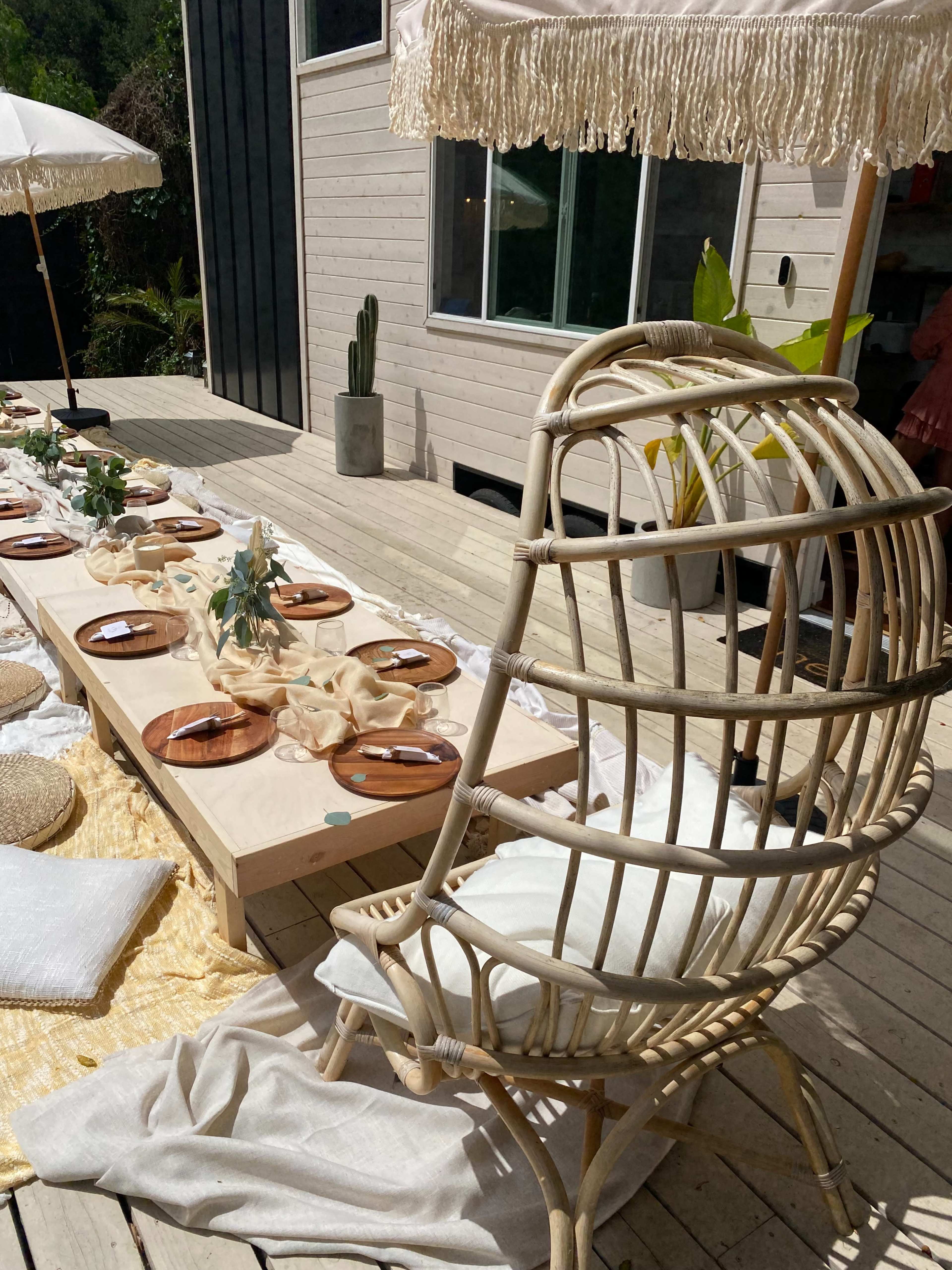 A spacious outdoor dining area features a long table set with plates and decor, accompanied by a large rattan chair and an umbrella, surrounded by greenery.