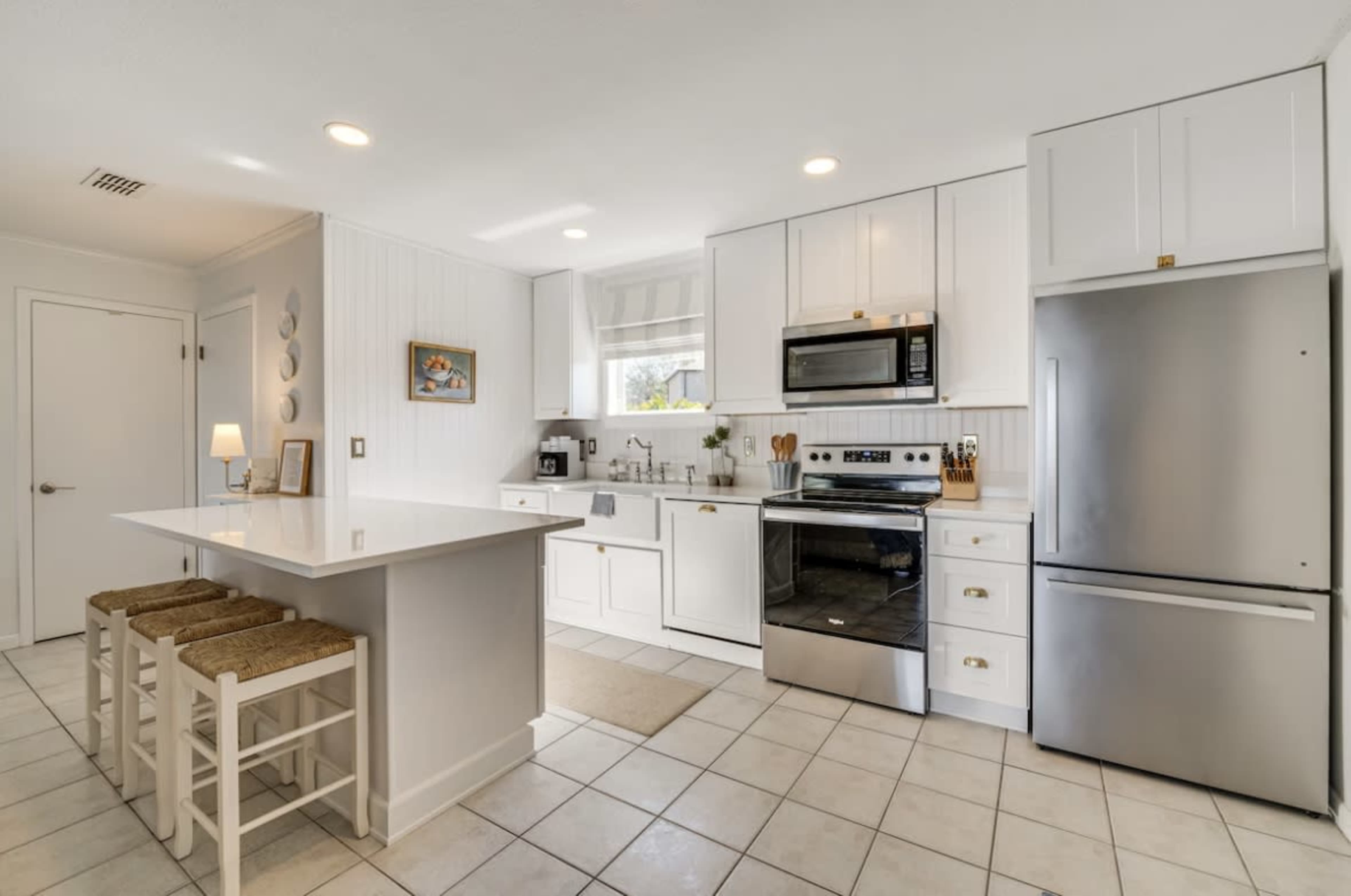 The image shows a modern kitchen with white cabinetry, stainless steel appliances, and a breakfast bar with two stools.