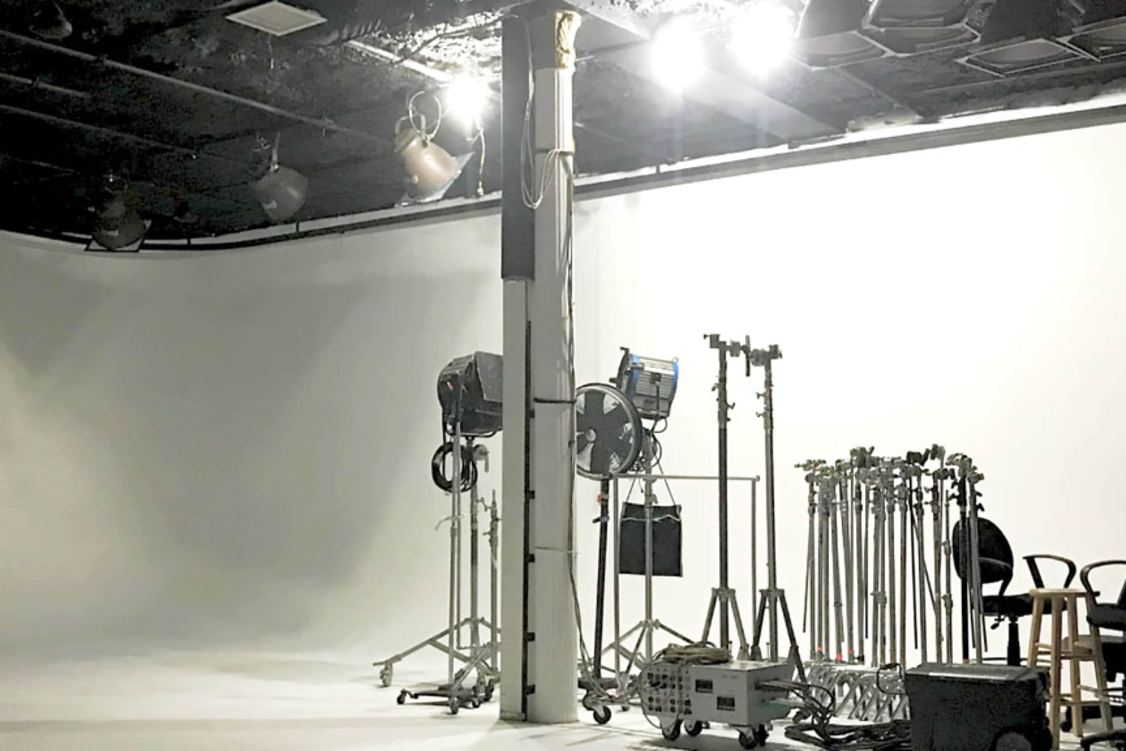A minimalistic photography studio features a white backdrop, lighting equipment, and various stands arranged around a central column.