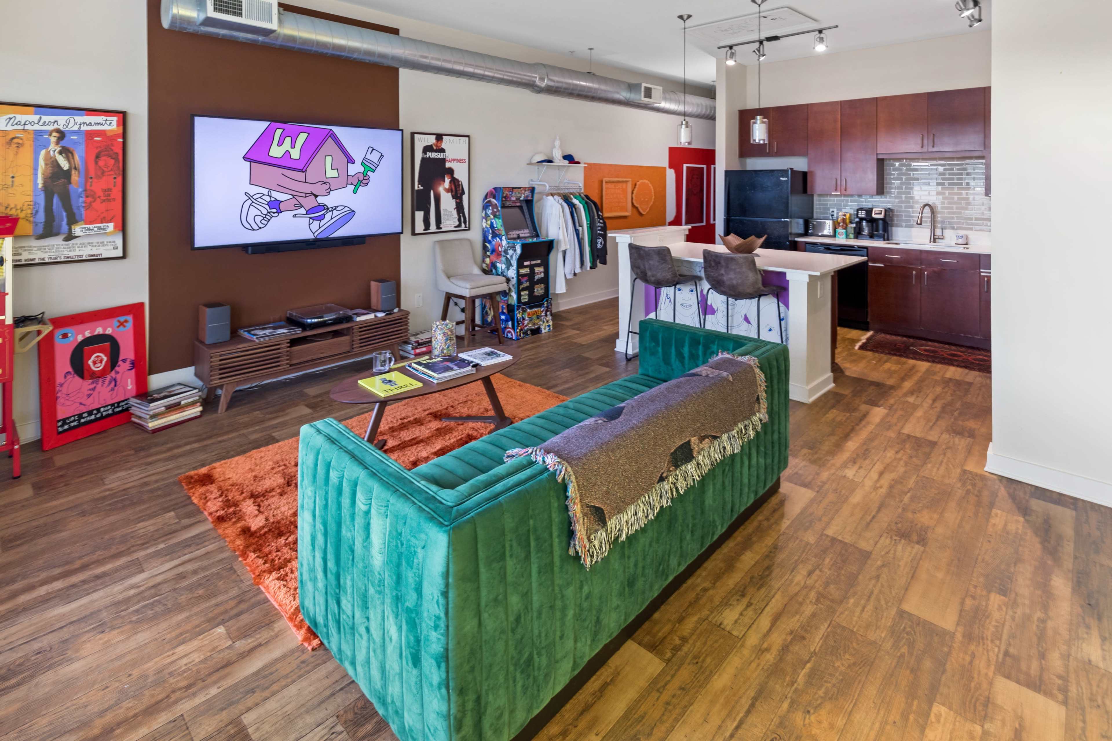 A modern living room with a green couch, a television displaying animated art, and an open kitchen area featuring dark wood cabinets and appliances.
