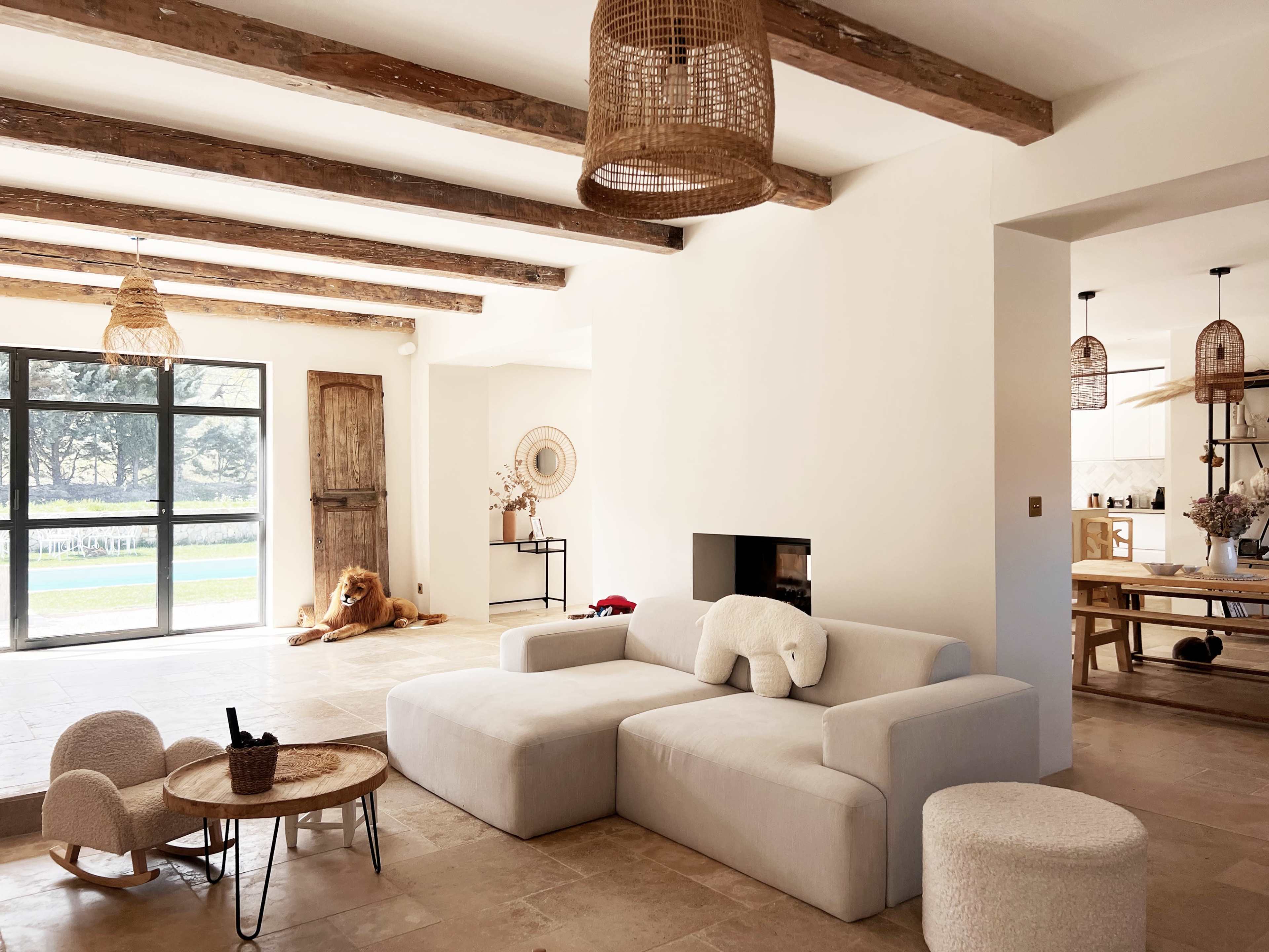 The image shows a modern, light-filled living room featuring a large beige sectional sofa, wooden beams on the ceiling, and a view of a sliding glass door leading to an outdoor space.