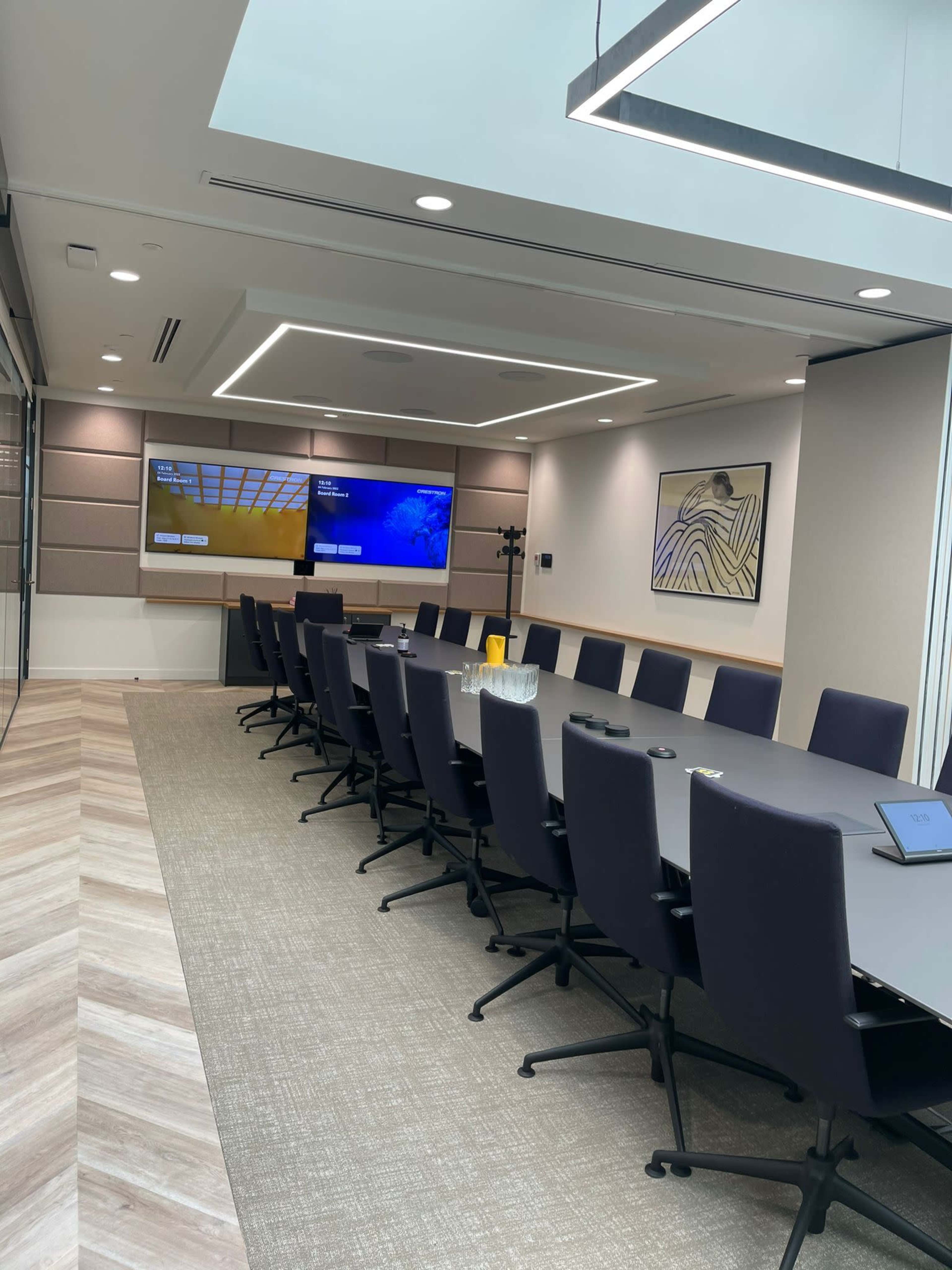 The image shows a modern conference room with a long table, black chairs, and two screens mounted on the wall.
