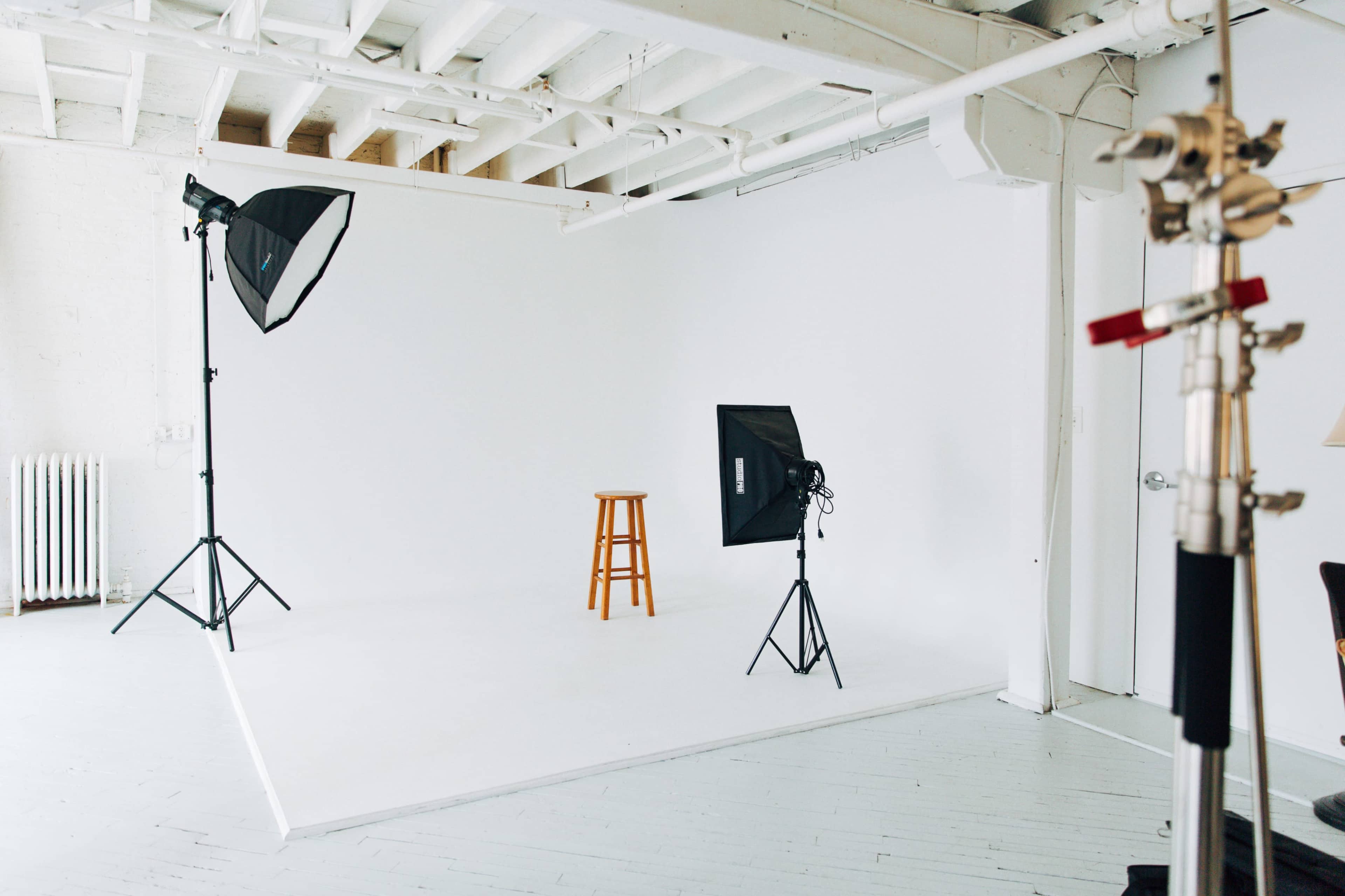 A photography studio features a white backdrop, a wooden stool in the center, and two softbox lights on stands.