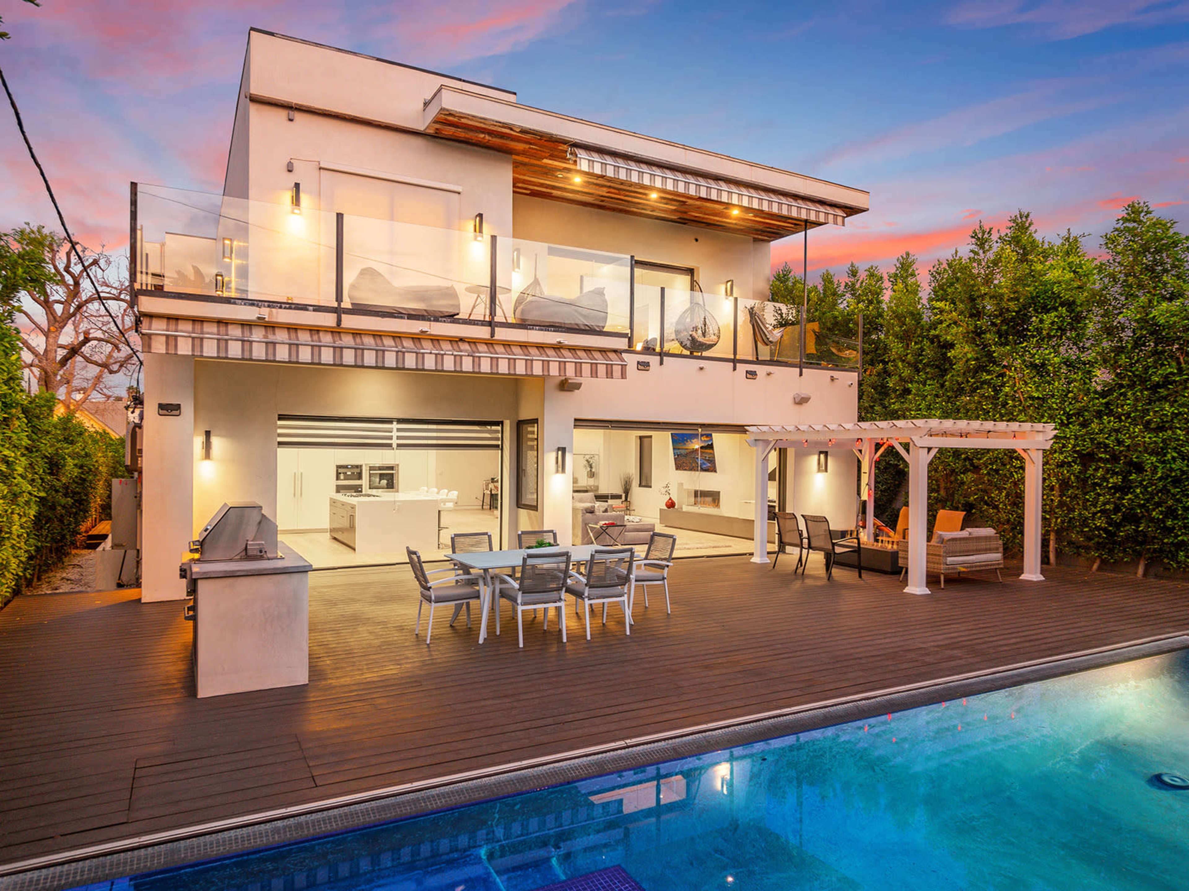 A modern two-story house with a patio, pool, and outdoor seating area is surrounded by greenery at sunset.