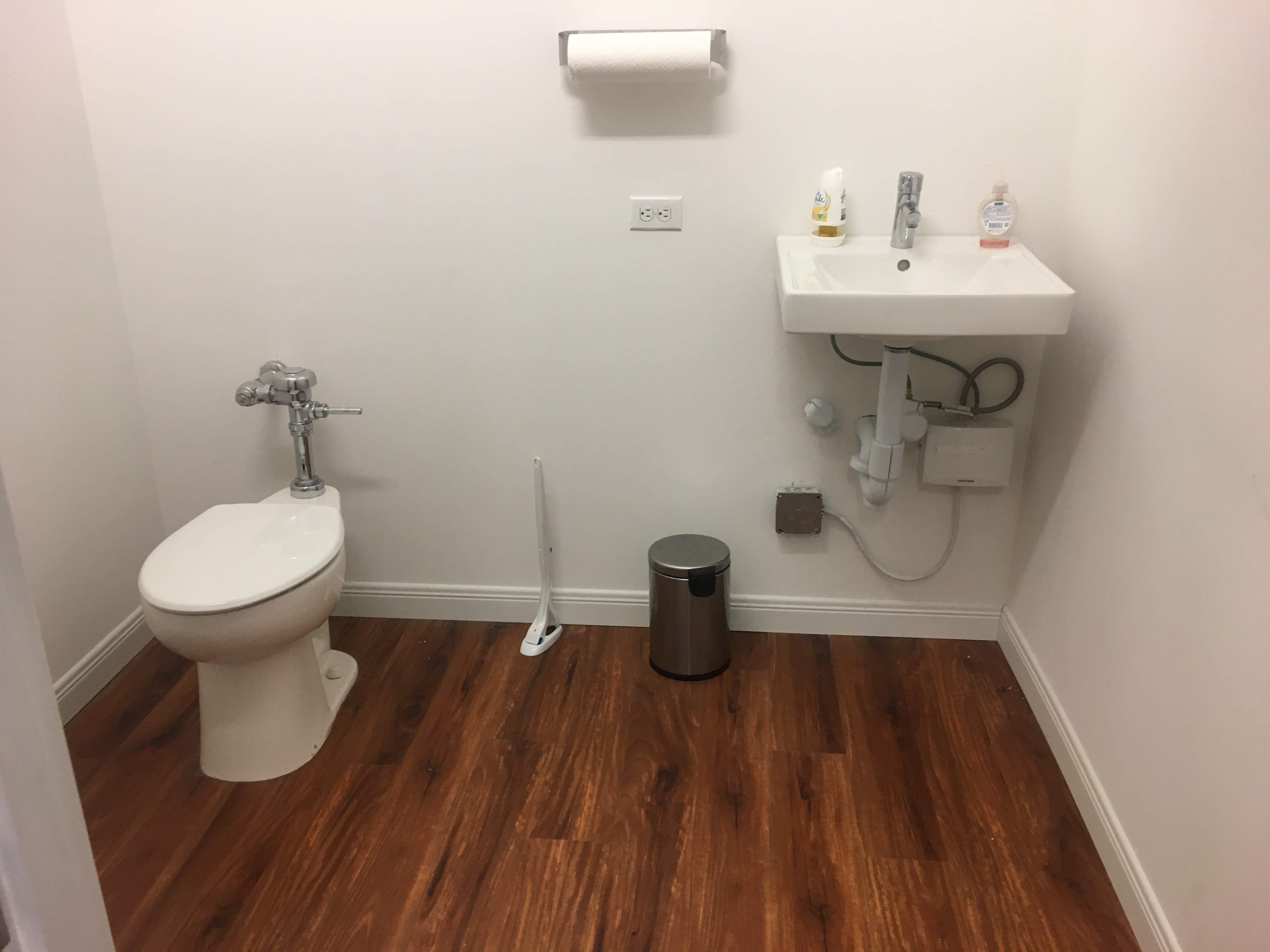 A small restroom with a toilet, a wall-mounted sink, a trash can, and a toilet brush on a wooden floor.