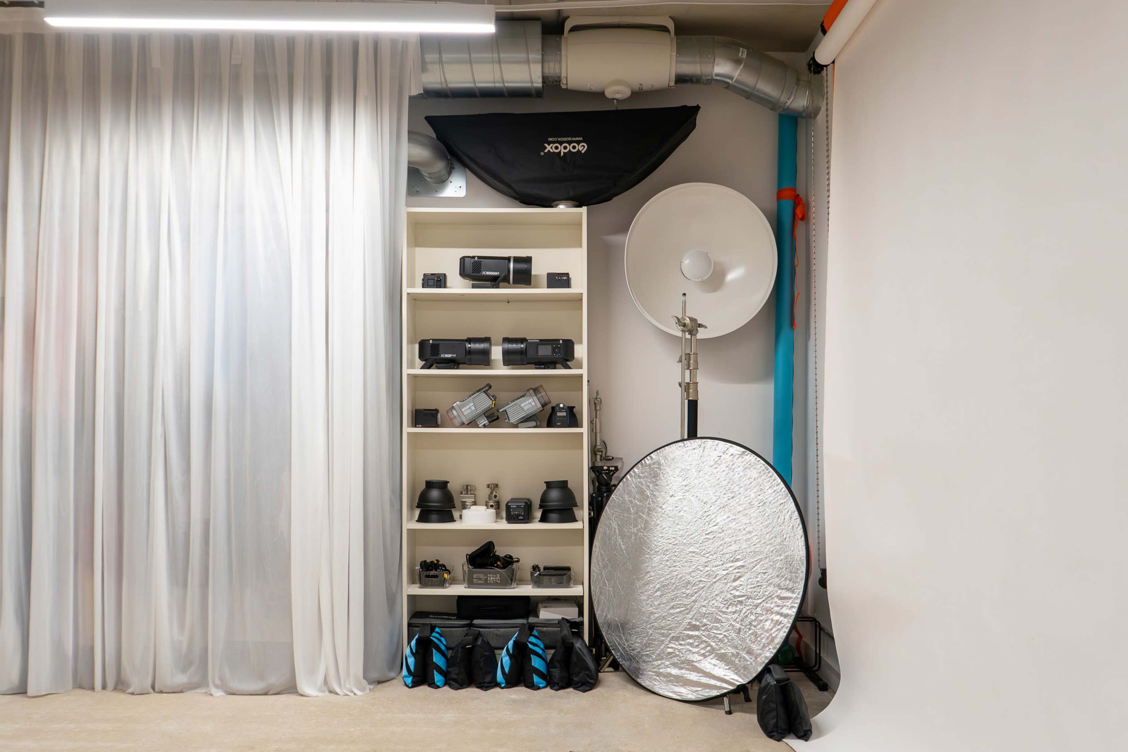 The image shows a photography studio setup with shelves displaying various camera equipment, softboxes, and reflectors, accompanied by a white backdrop and curtains.
