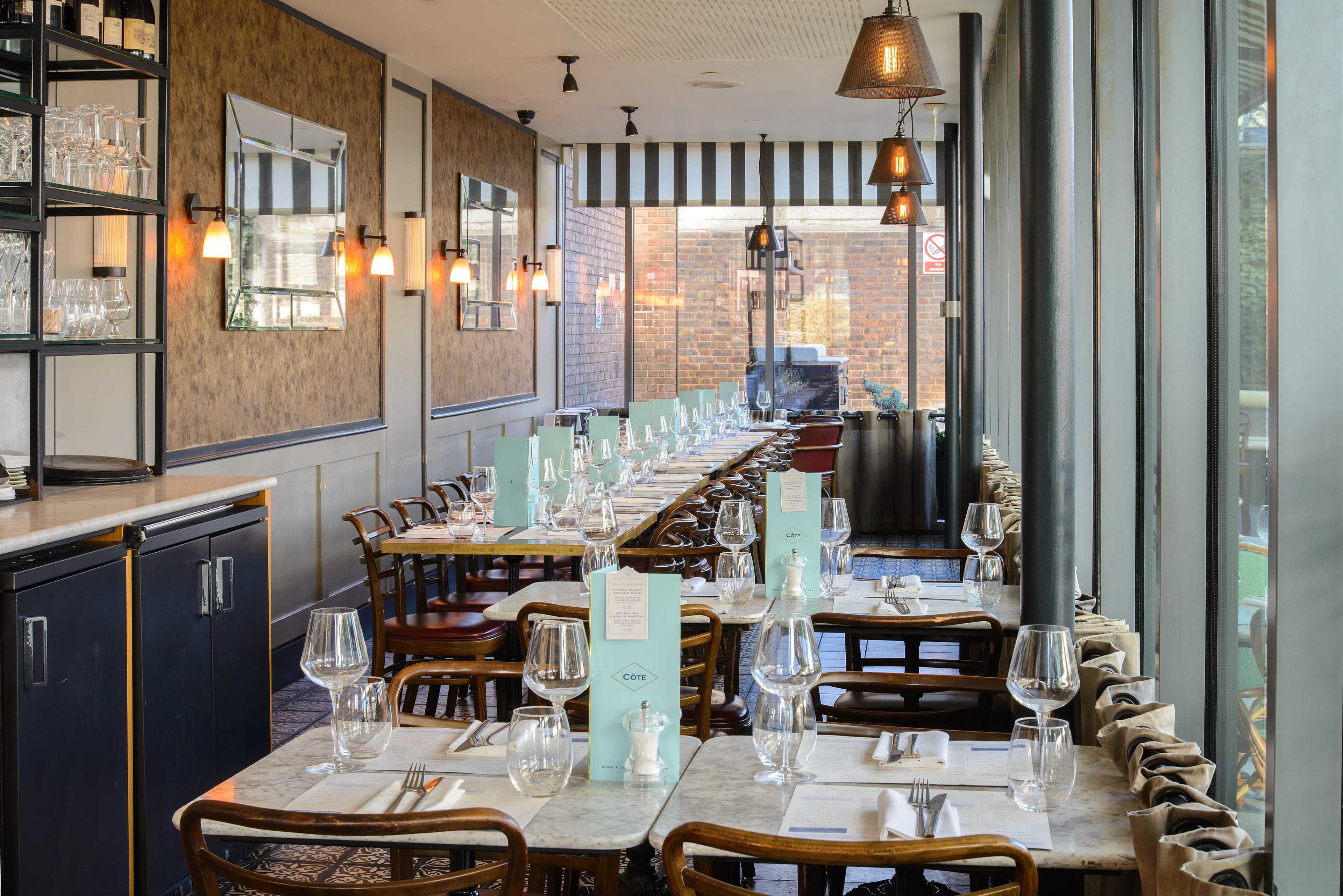 The image shows a modern restaurant interior with a row of tables set with glassware and menus, flanked by large windows and decorative lighting.