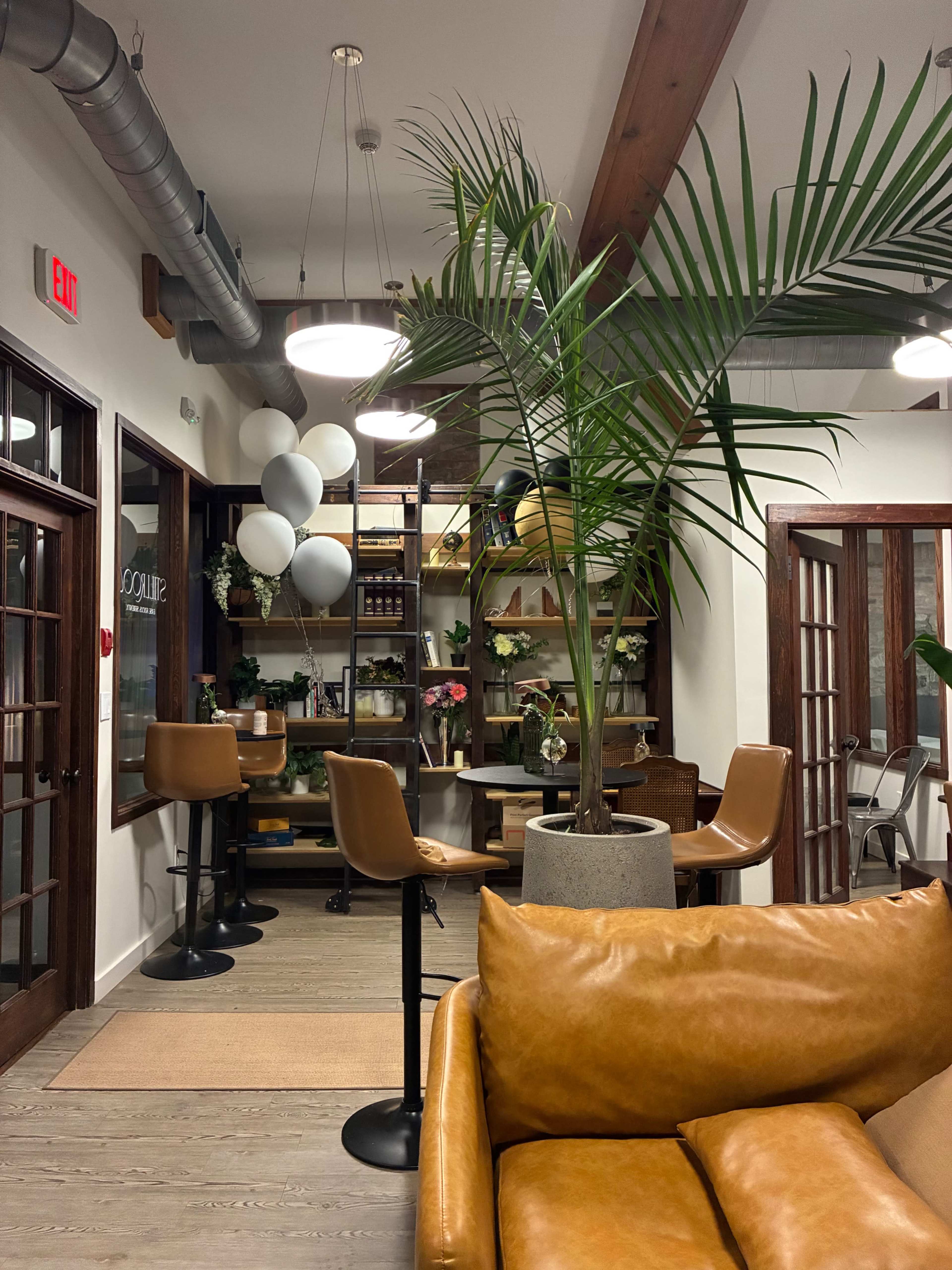 The image shows a stylish interior space with a brown leather sofa, tall potted palm, and a seating area featuring bar stools and a table, surrounded by shelves decorated with plants and various items.