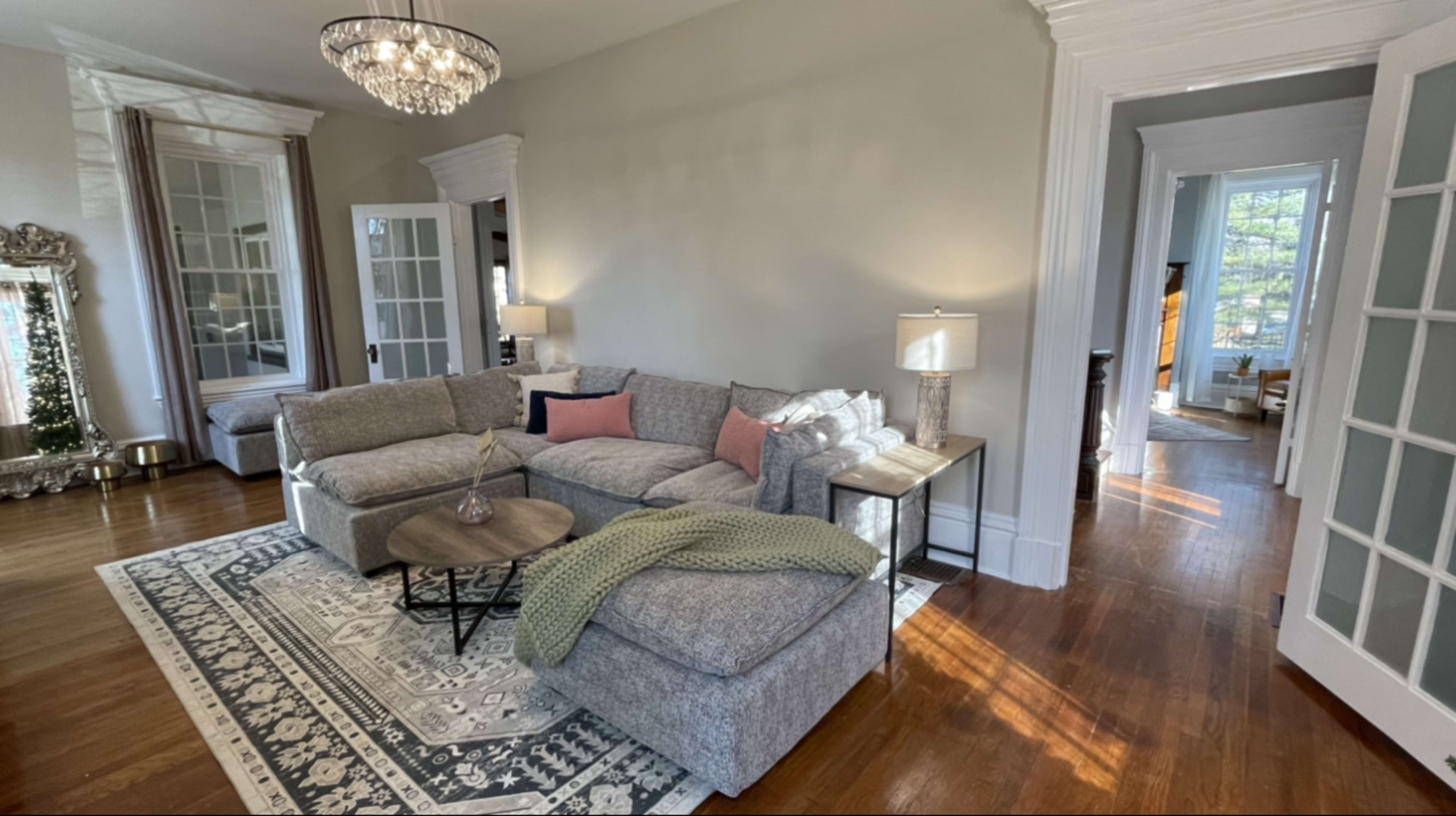 A well-lit living room featuring a grey sectional sofa, a round coffee table, and a large mirror, with French doors leading to a bright outdoor area.