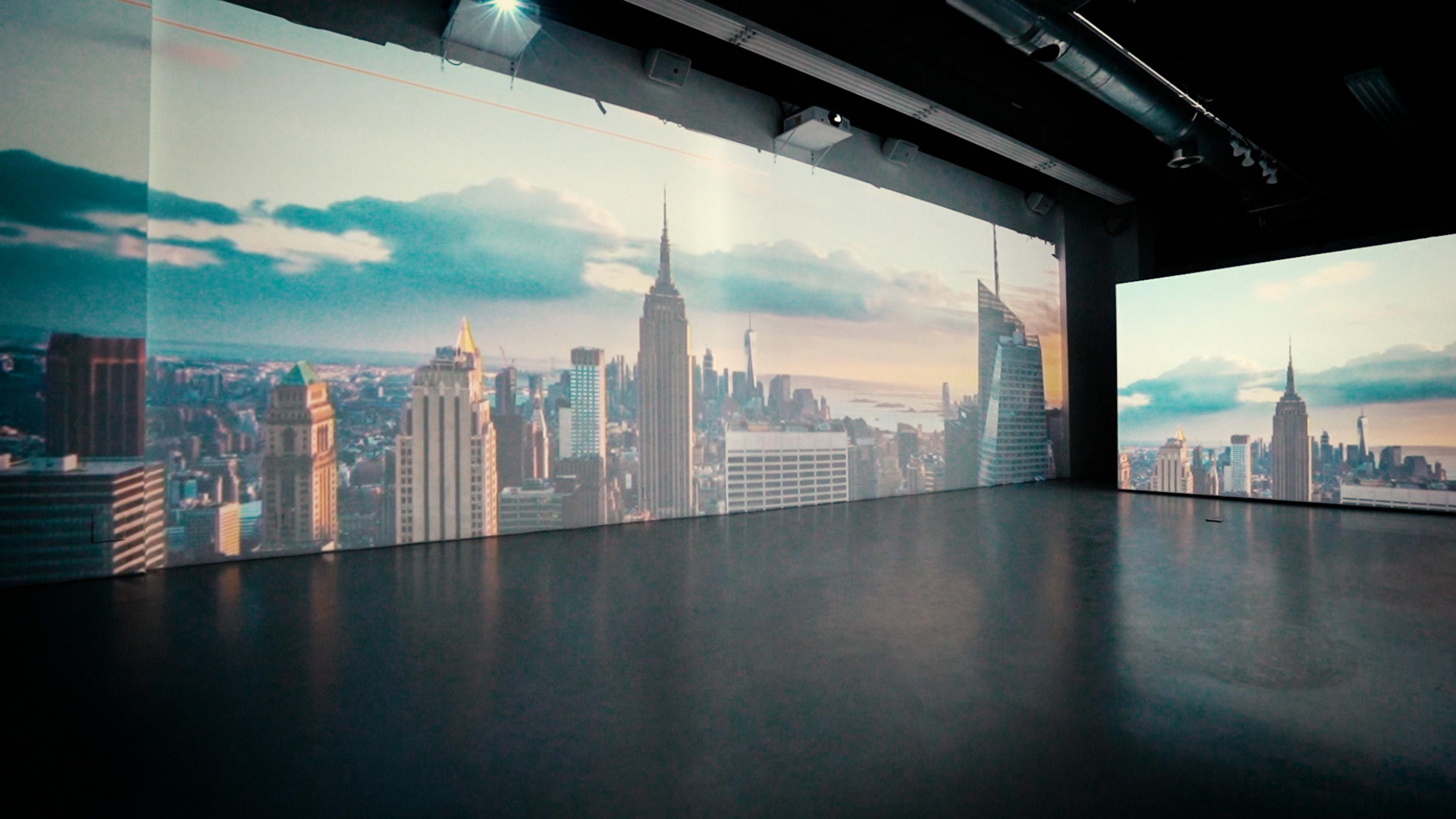 A spacious gallery with large projections of a New York City skyline on the walls.