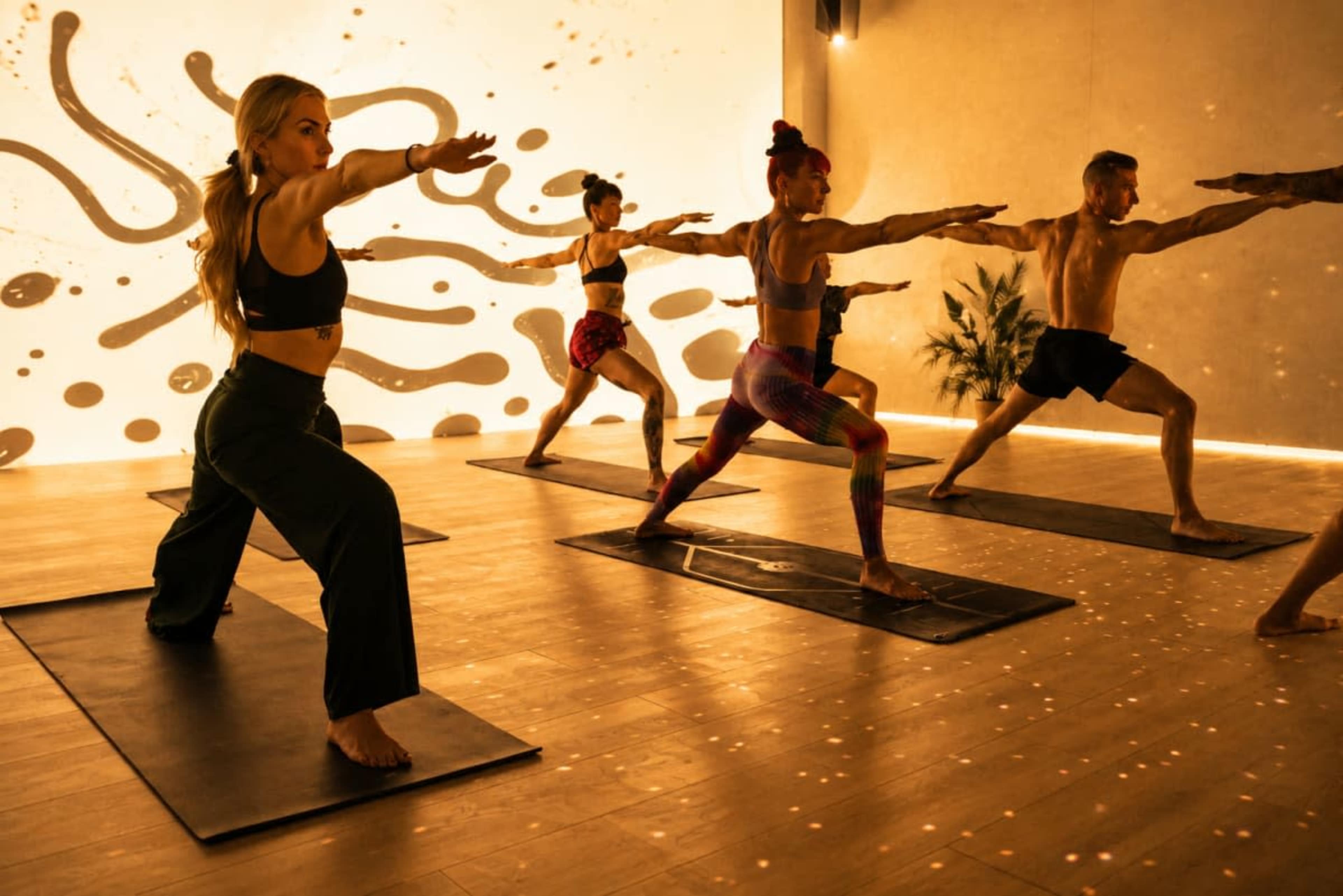 A group of individuals is practicing yoga in a brightly lit studio with abstract wall designs, each person in a different pose on their mats.