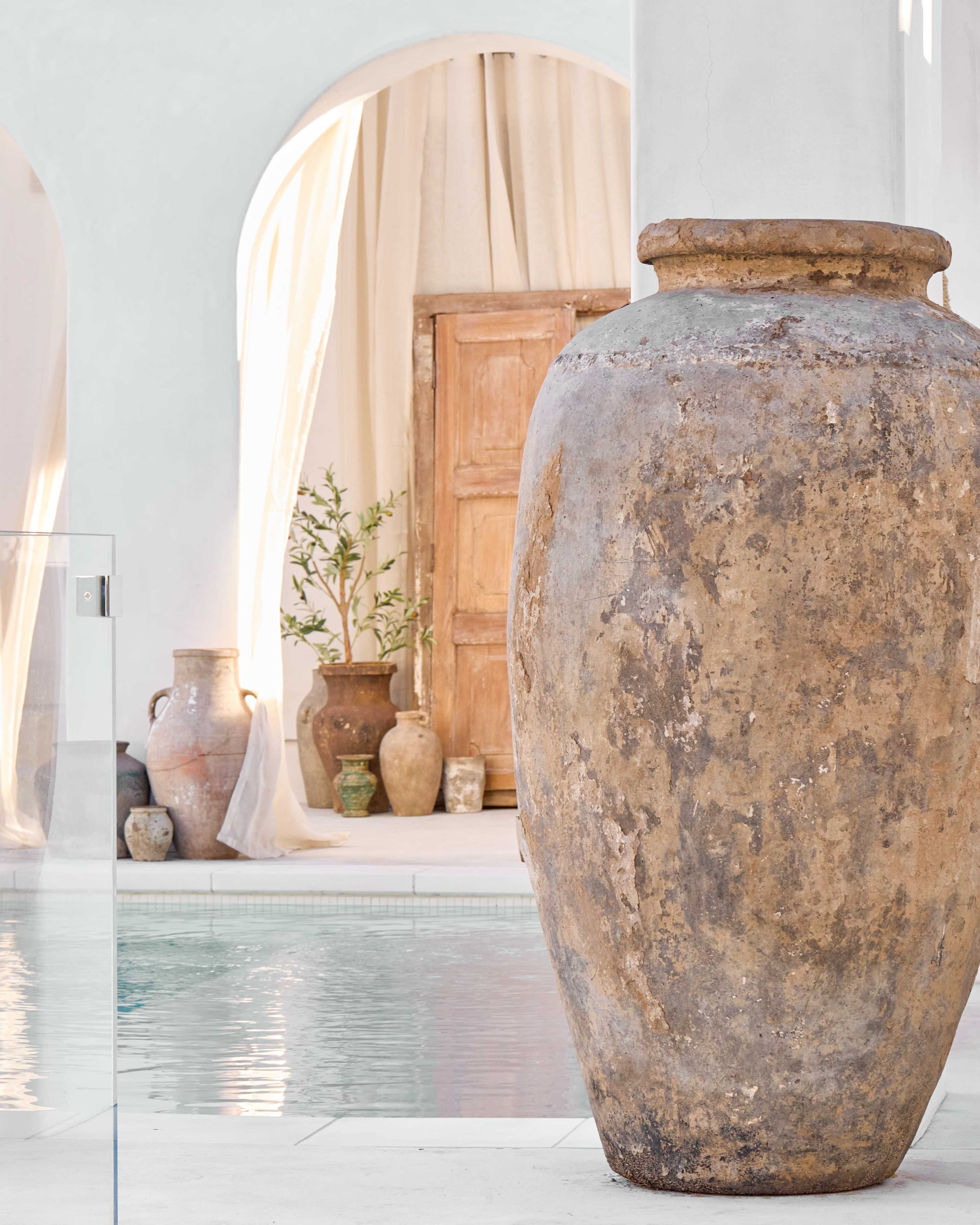The image features a large, weathered pottery vessel beside a swimming pool, with an open doorway and additional smaller pottery pieces in the background.