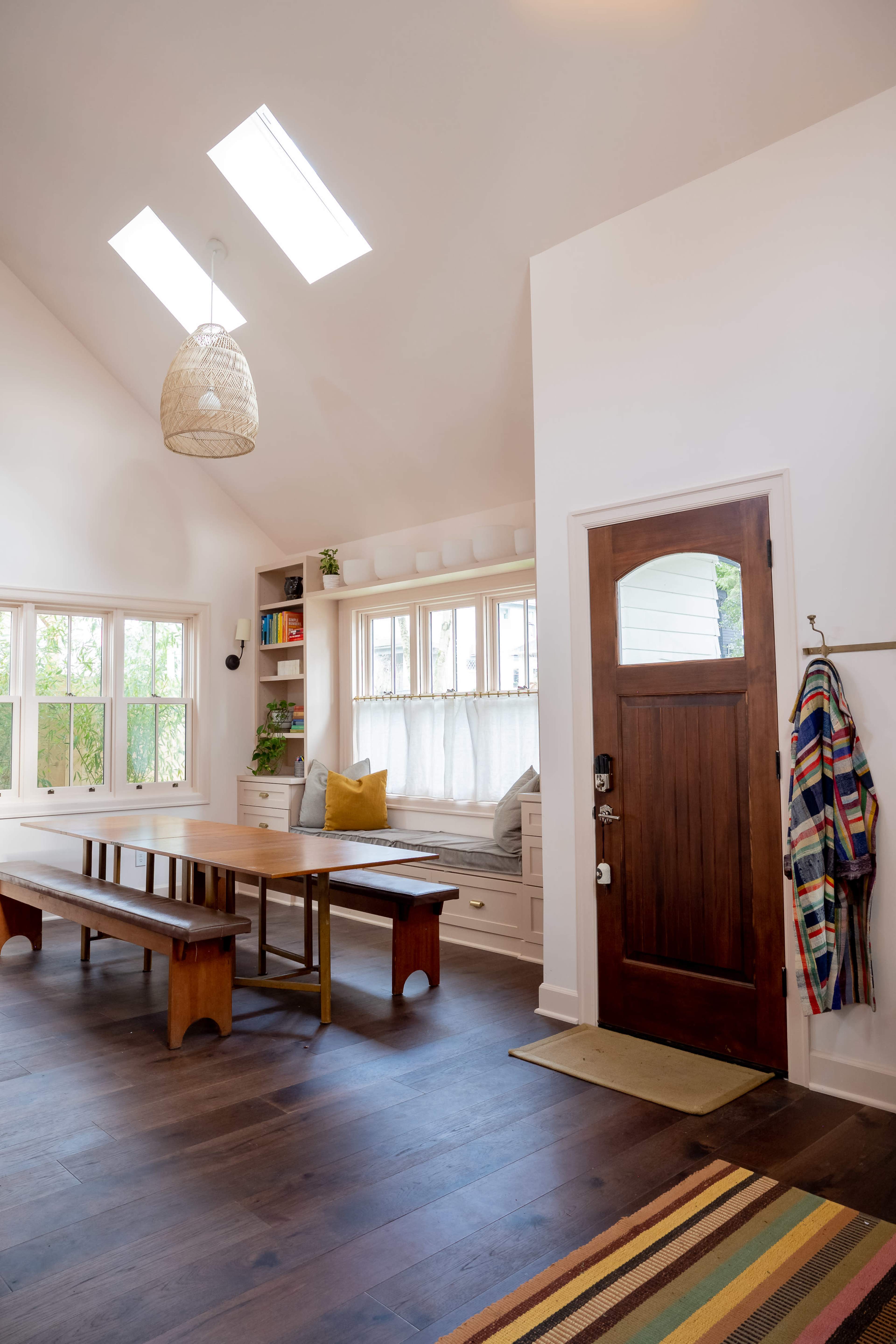 A bright, modern entryway with a wooden front door, a dining table, and a cozy reading nook by the windows.