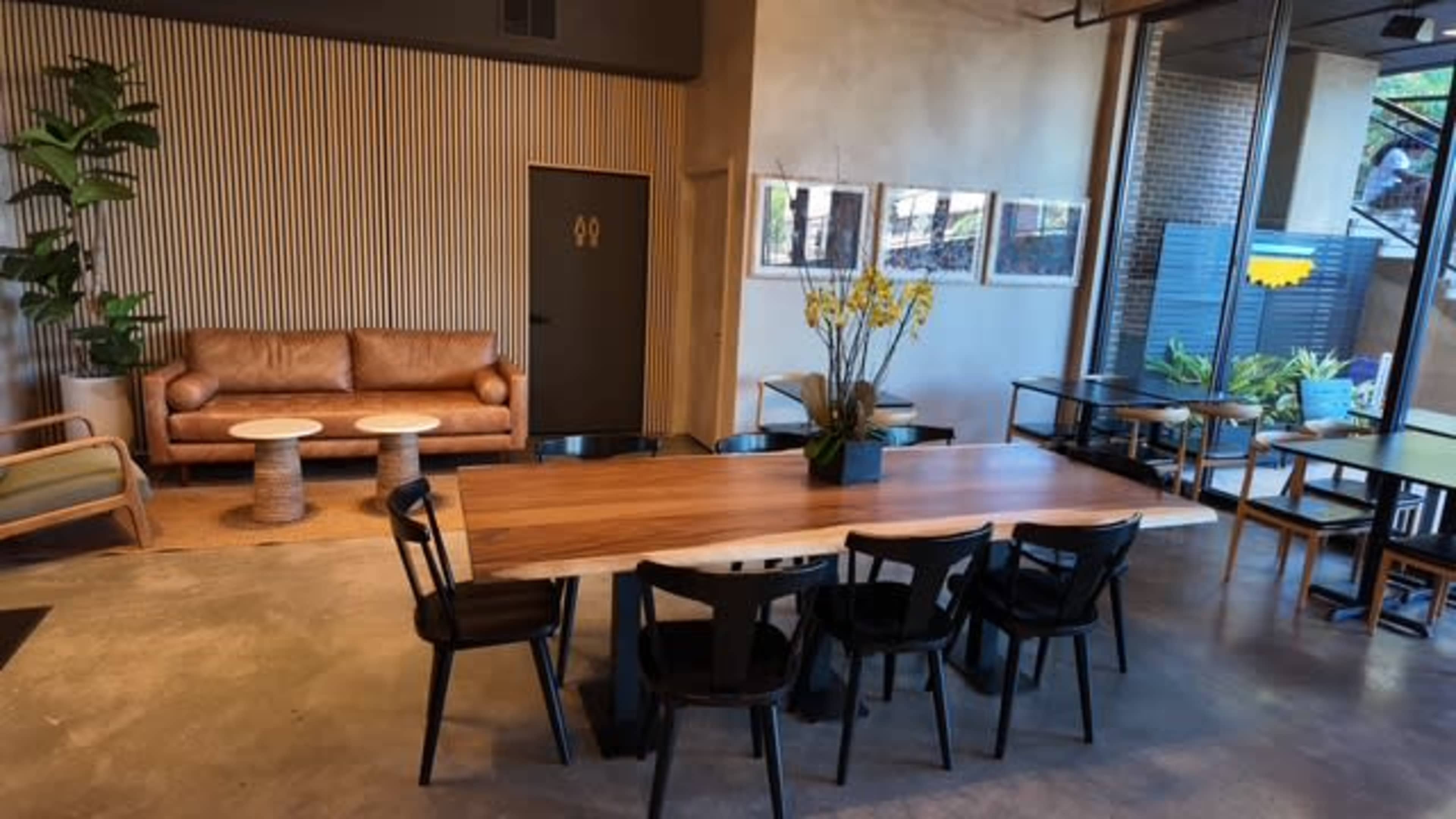 The image shows a modern interior space featuring a wooden dining table surrounded by black chairs, a brown leather couch, and potted plants.