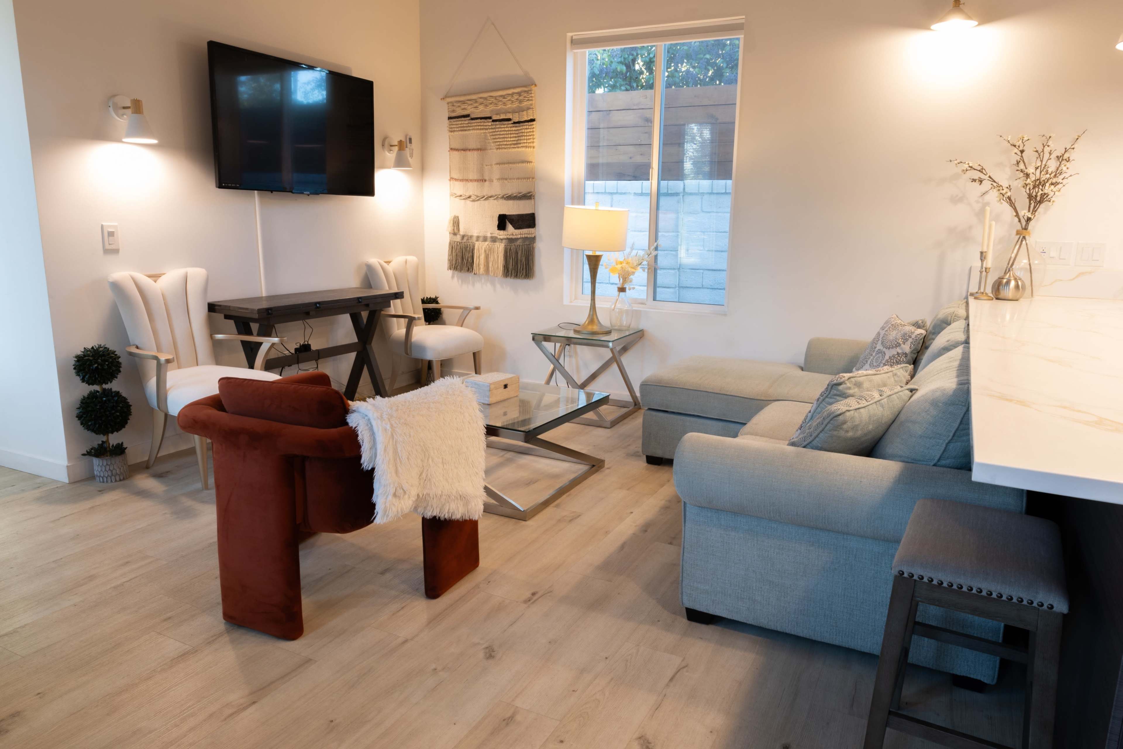 The image shows a modern living area with a television mounted on the wall, seating arrangements that include a light gray sofa, two chairs, and a small table, along with a side table and a decorative lamp.