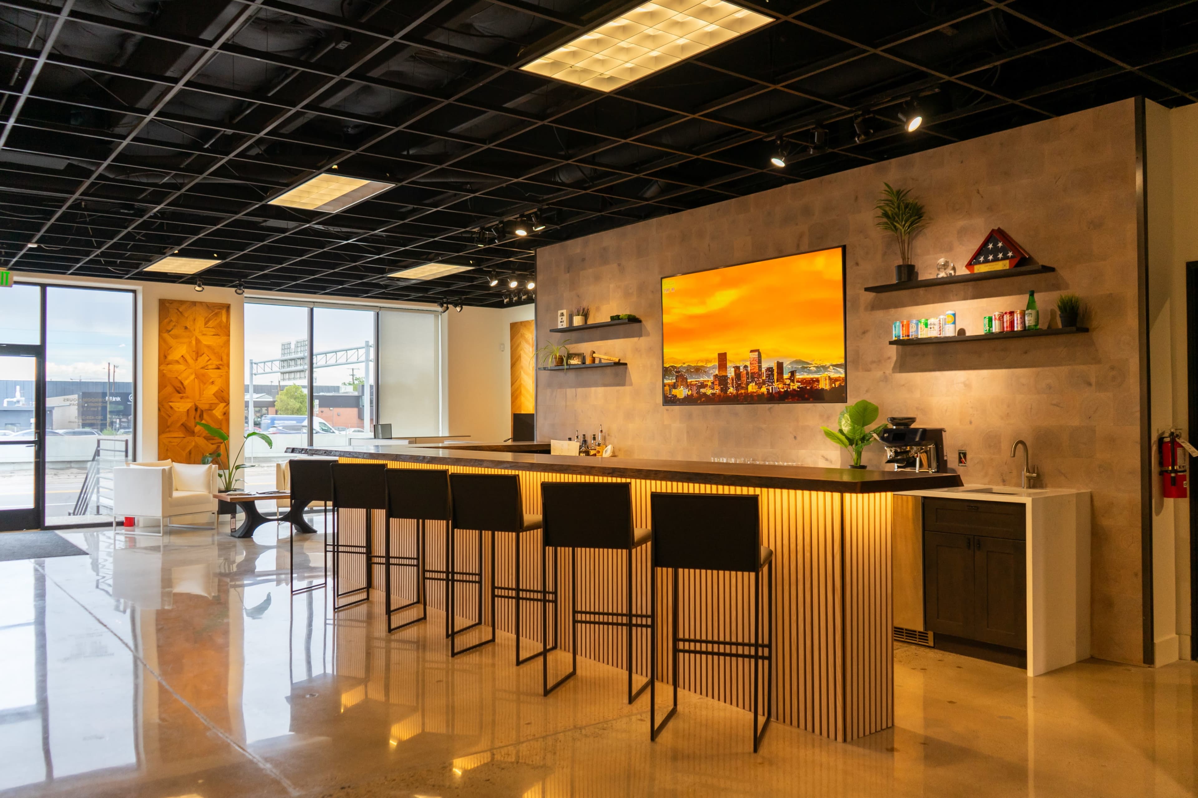 The image shows a modern kitchen area with a long bar counter, high stools, and a large wall-mounted television displaying a cityscape.