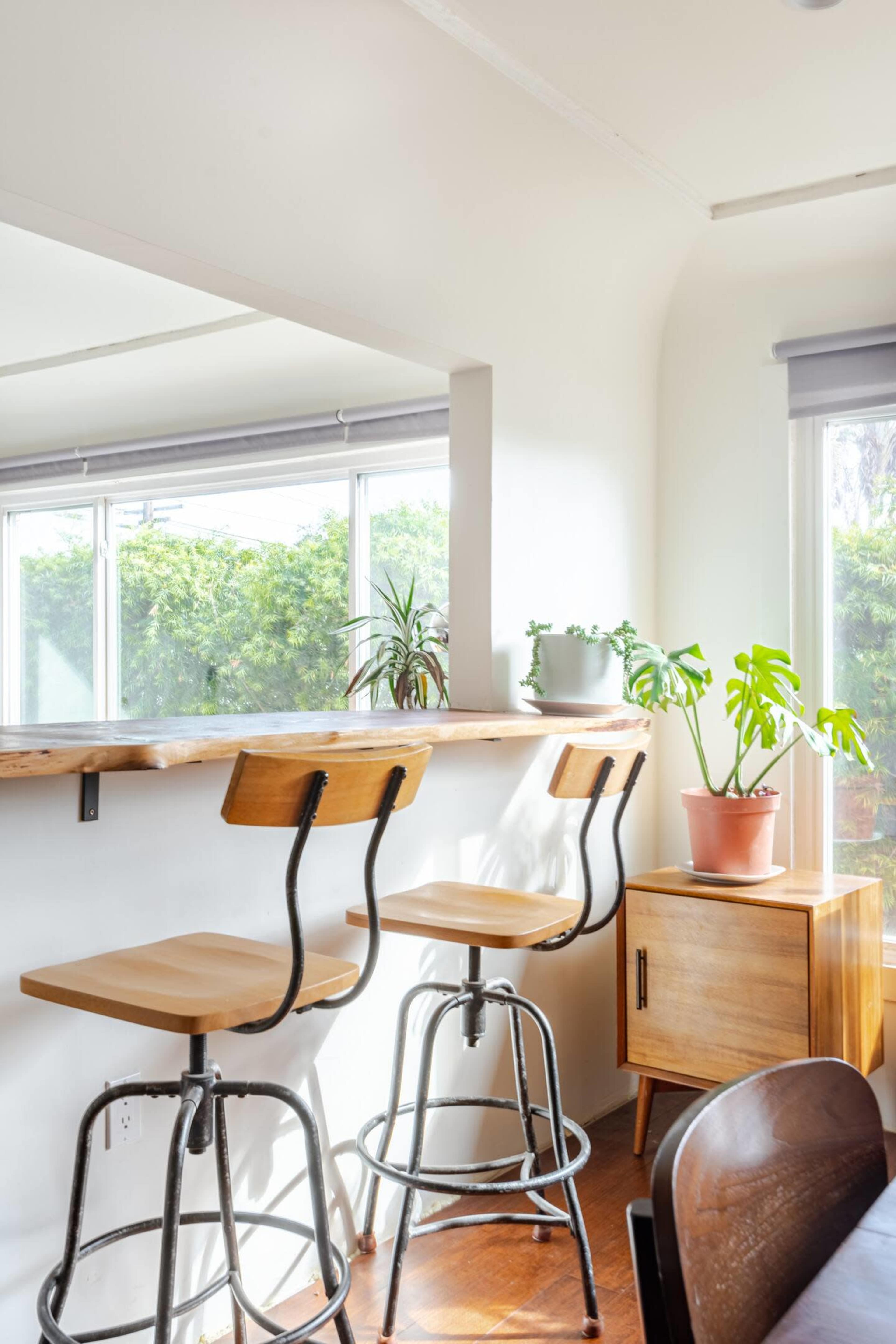 A bright corner features two wooden bar stools at a simple counter, with potted plants and a wooden cabinet nearby.