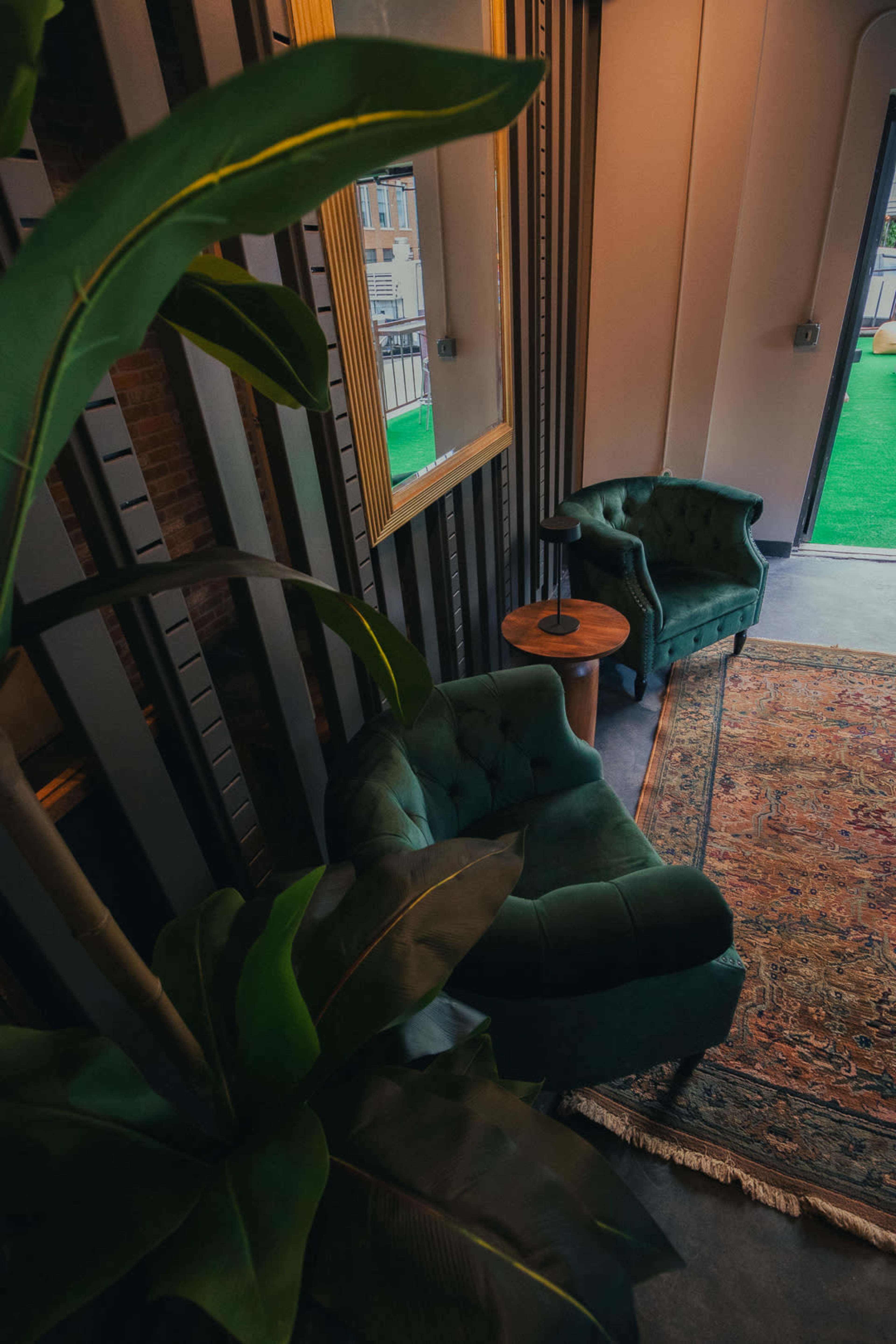 A cozy corner features two green, tufted armchairs beside a wooden table and a large plant, with a decorative rug on the floor and a mirror on the wall.