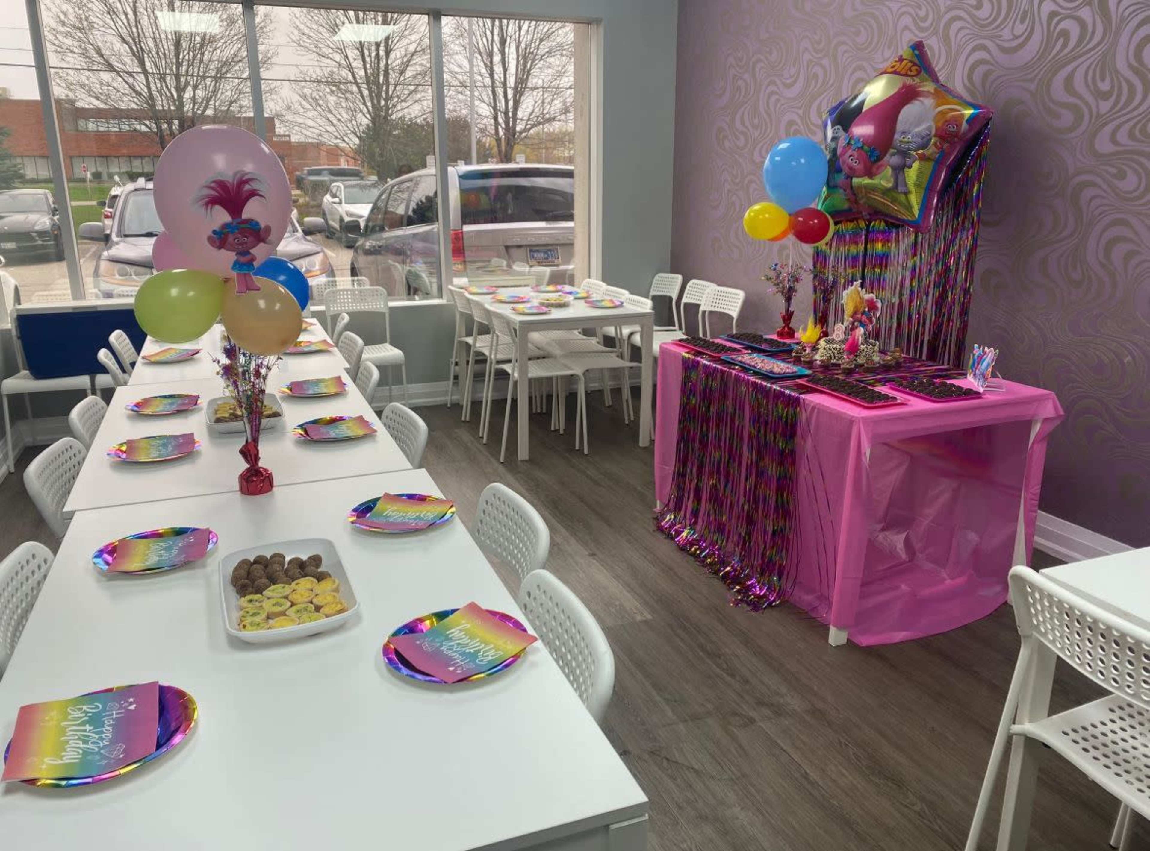 A bright birthday party setup features a long table with colorful plates and a dessert table decorated with balloons and a pink tablecloth.