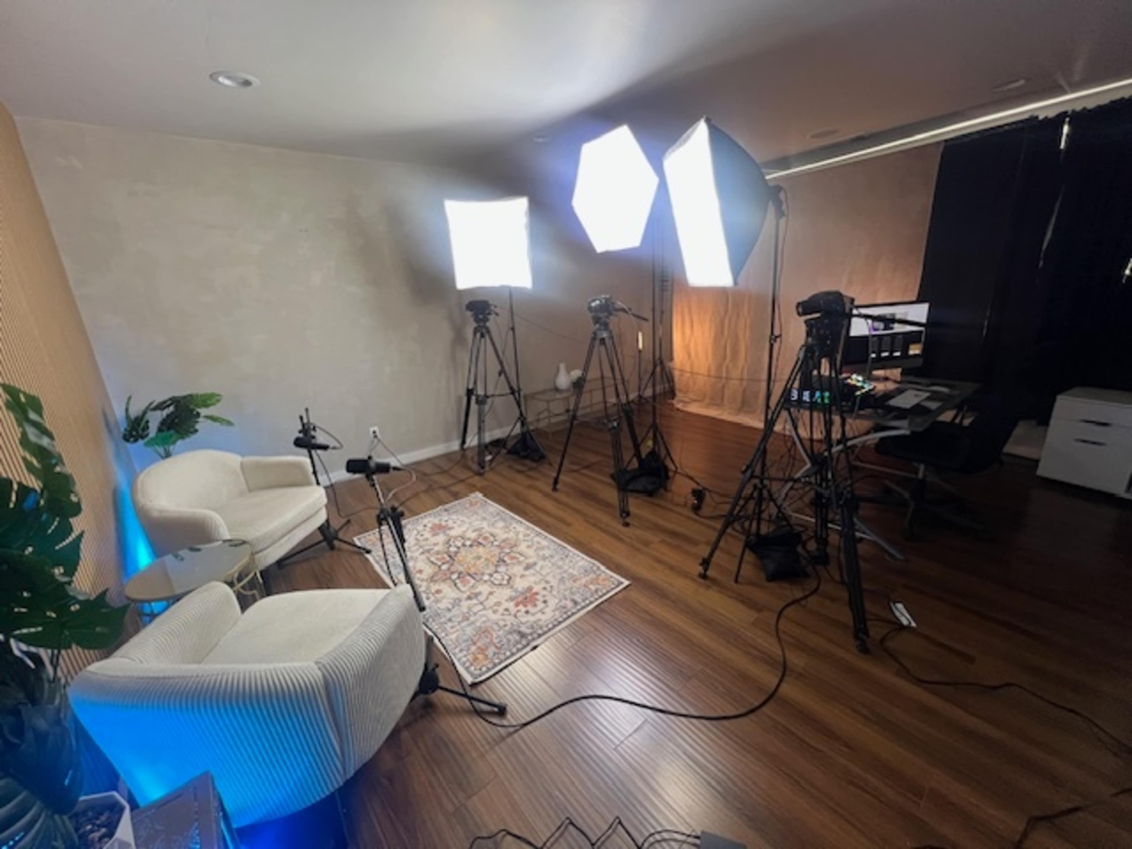 The image shows a well-lit studio setup featuring two white chairs, multiple tripods with lights, and a desk with devices in a room with wooden flooring.