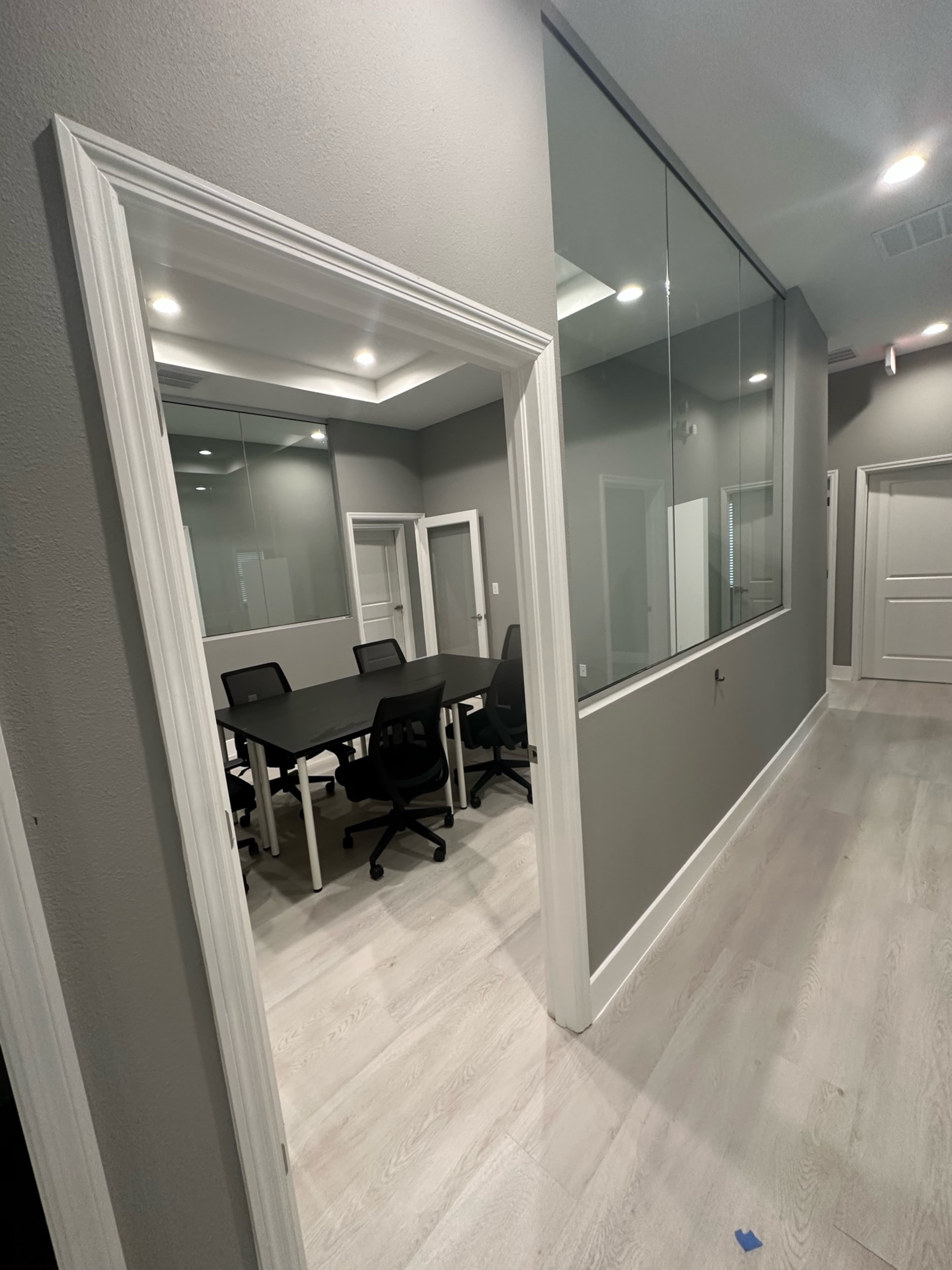 The image shows a modern office corridor leading to a conference room with a large table and several chairs, visible through a glass wall.