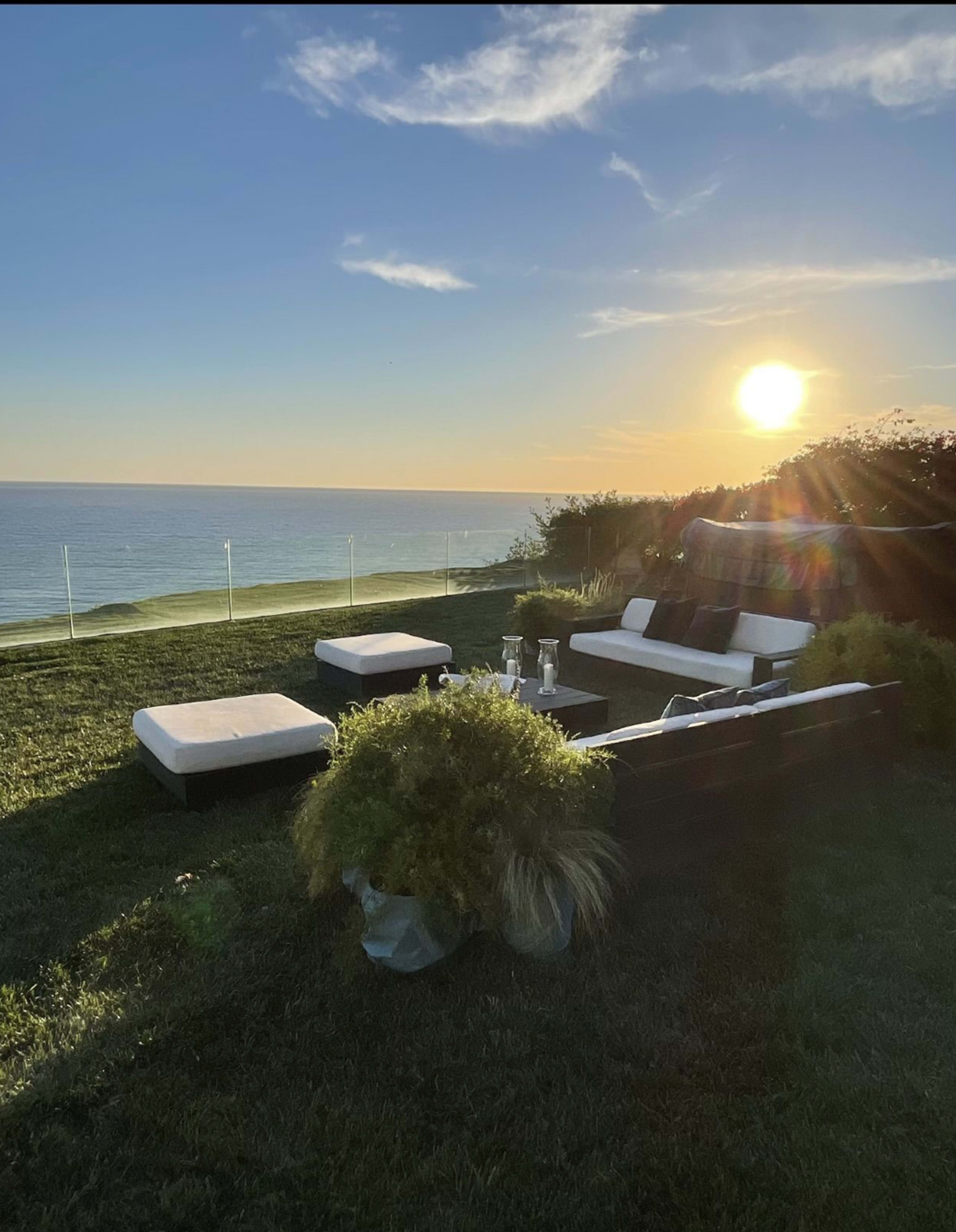 A modern outdoor seating area with white cushions overlooks the ocean at sunset.