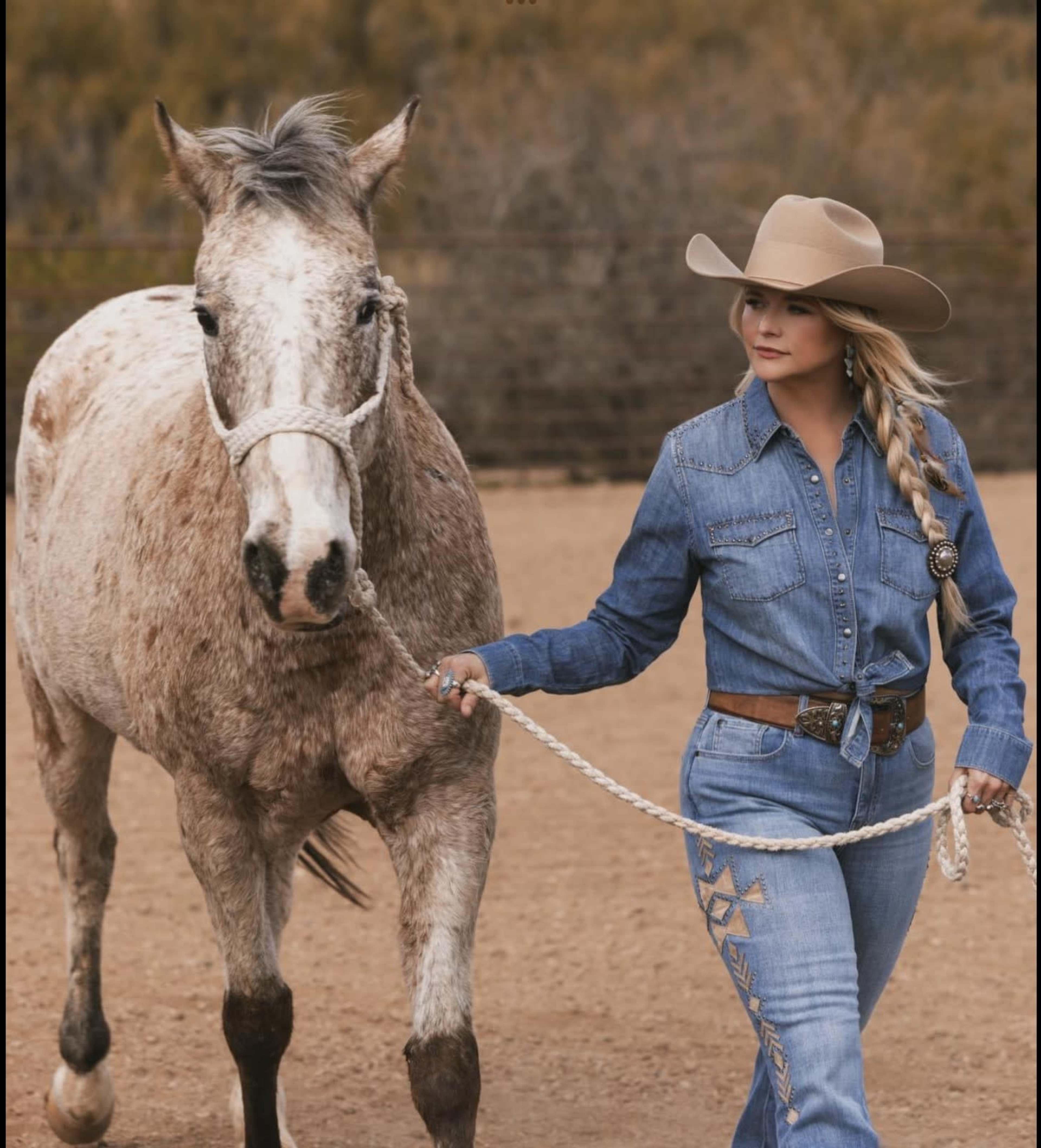 A person in a denim outfit walks alongside a gray horse in an outdoor arena.