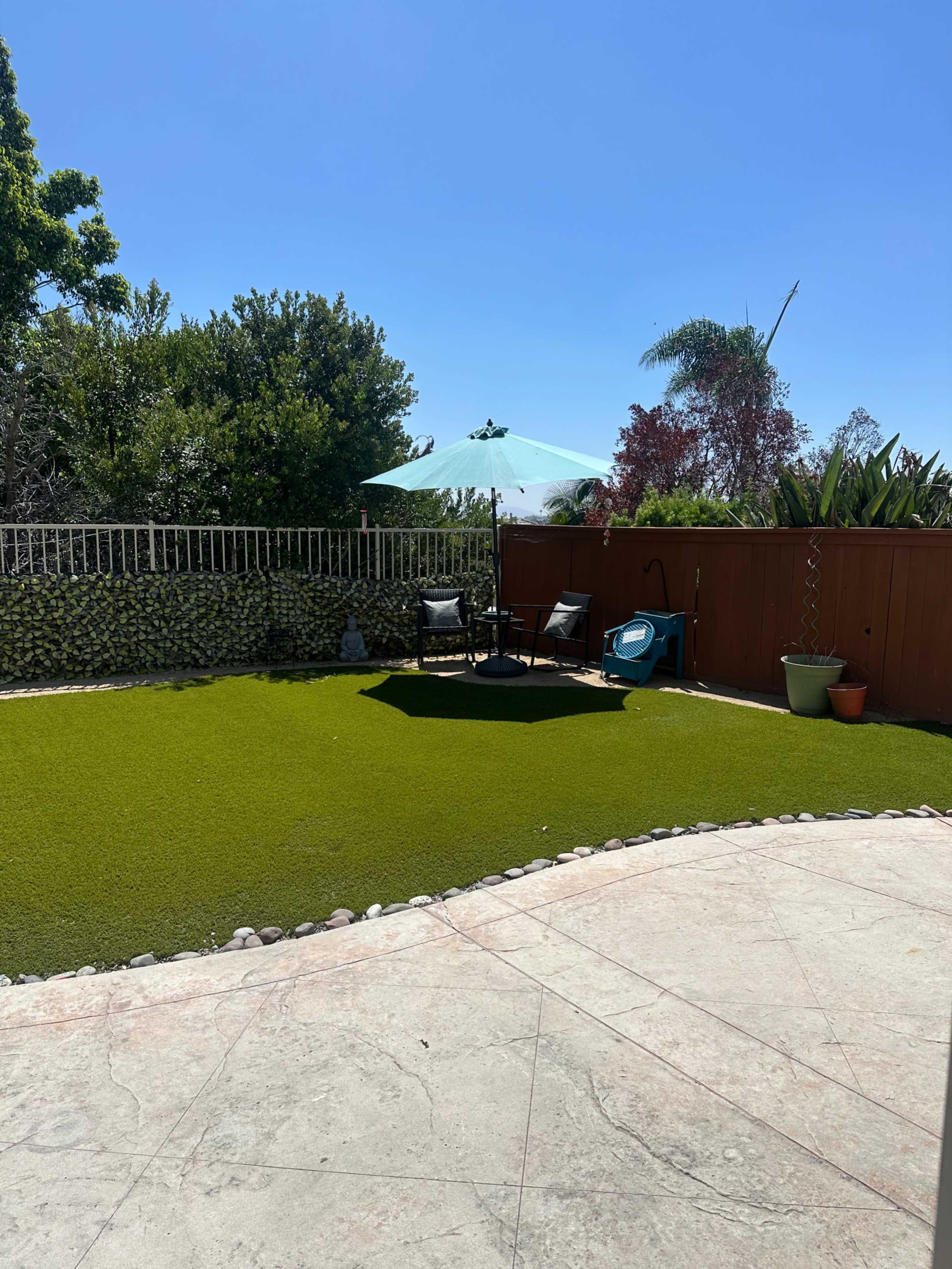 A well-maintained backyard featuring artificial grass, a patio area with stone borders, a large umbrella over a table and chairs, and various plants along the perimeter.