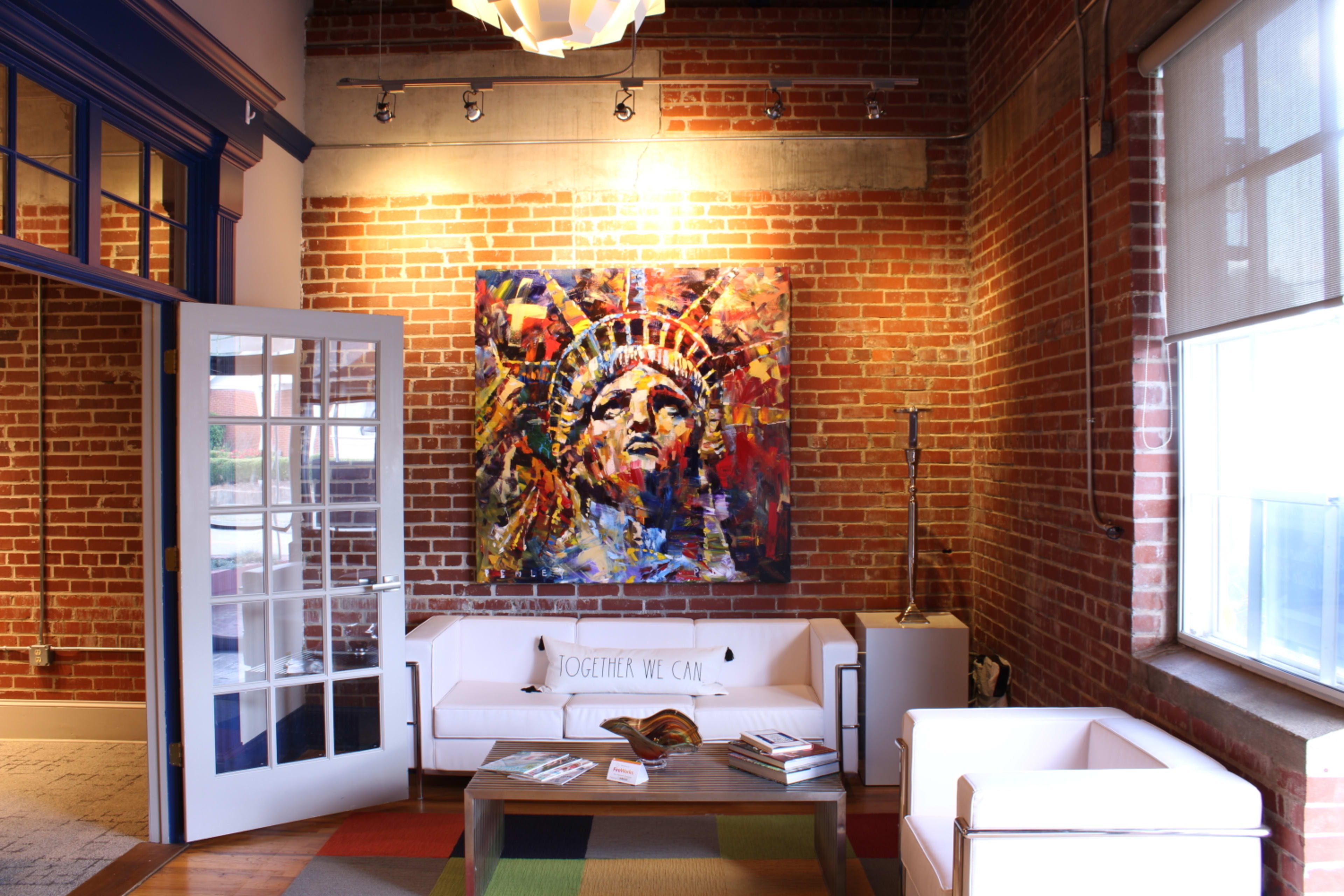The image shows a modern living area with exposed brick walls, white couches, a coffee table, and a colorful painting of the Statue of Liberty hanging above.