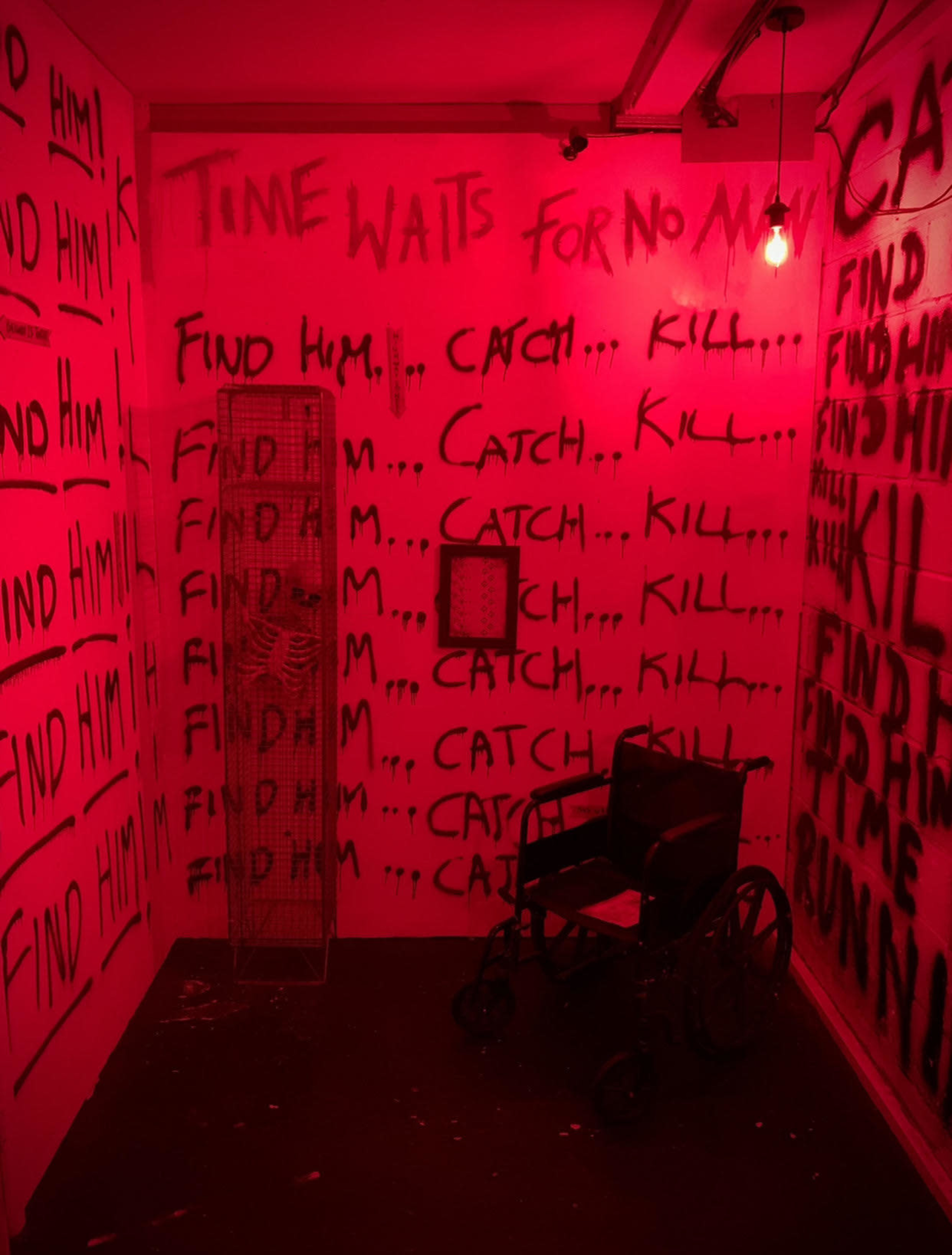 A dimly lit room features red walls covered in graffiti that reads "FIND HIM... CATCH... KILL..." alongside a wheelchair and a wire cage.