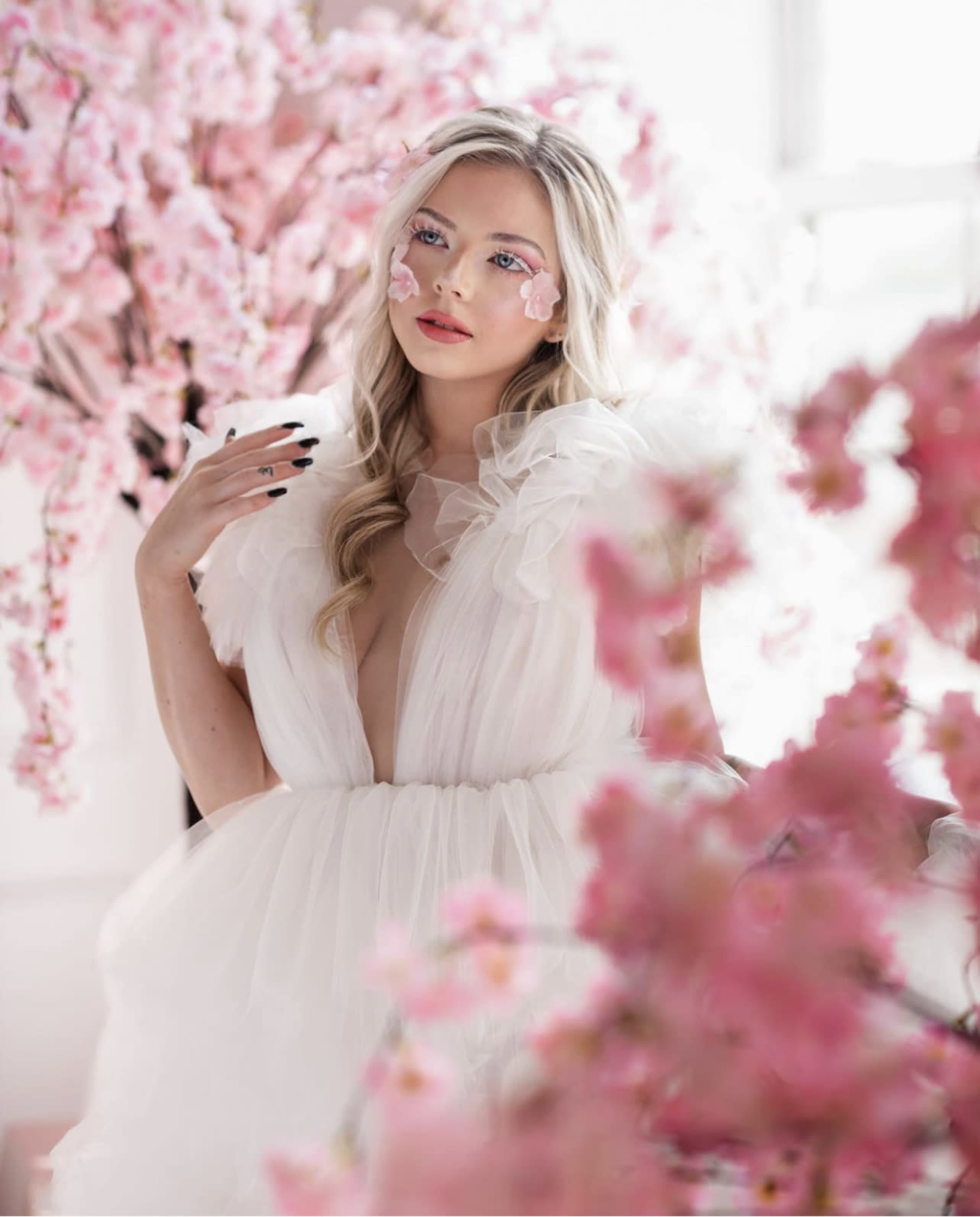 A woman in a white dress stands among pink flowering trees, with floral decorations on her face.