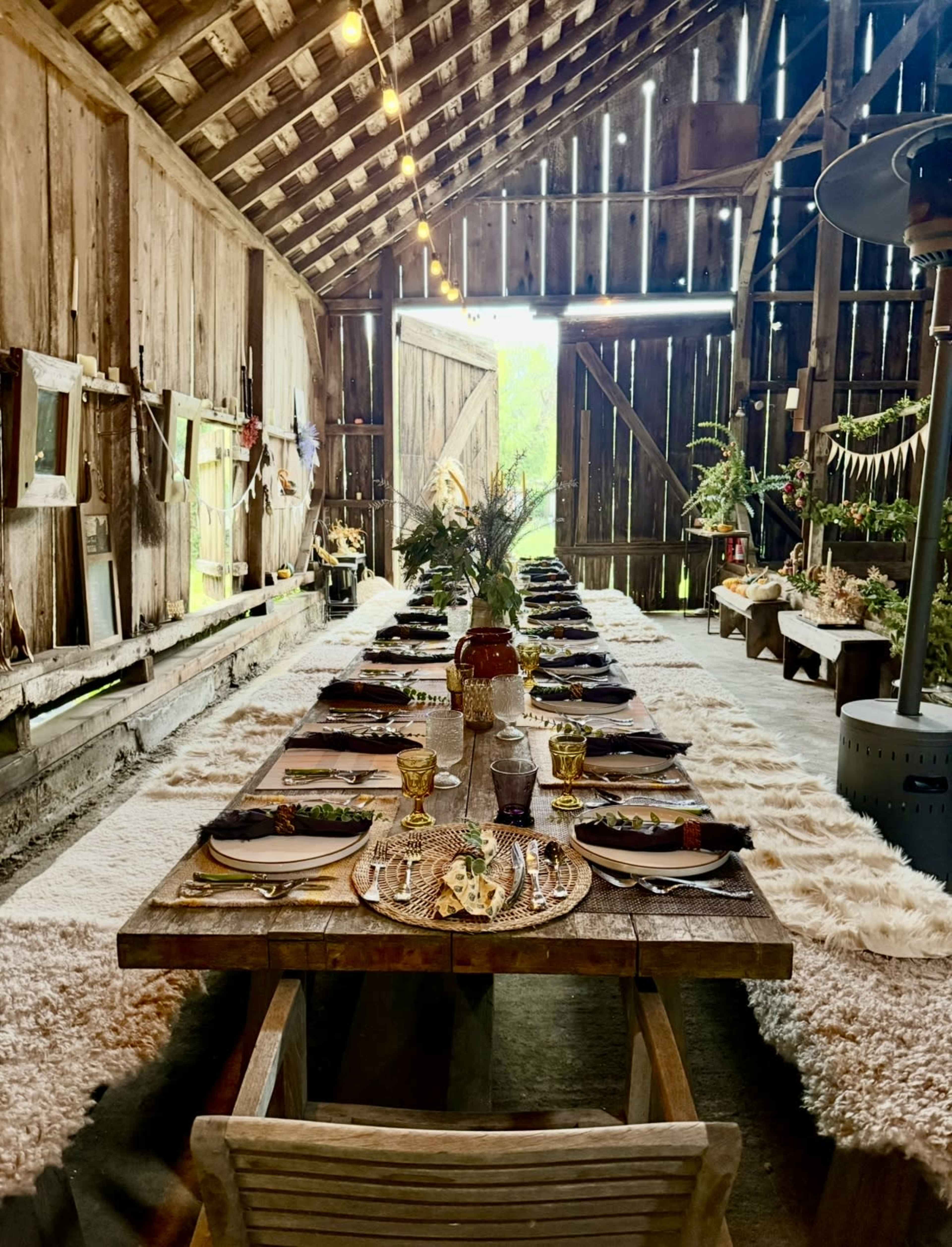 A long wooden table is set for a meal in a rustic barn, with neatly arranged plates and decor, and a soft rug covering the floor.