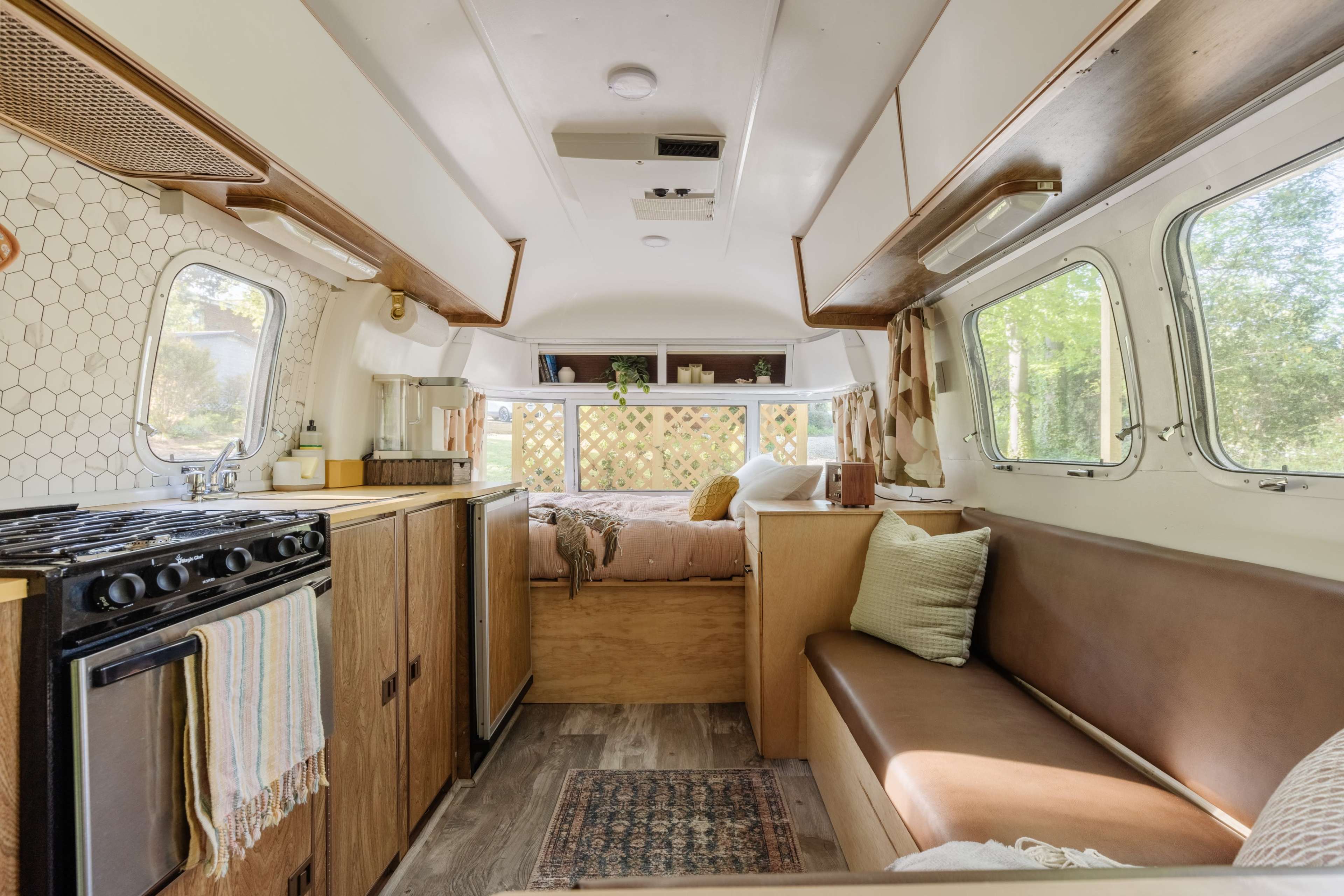 The image shows the interior of a recreational vehicle, featuring a kitchen area with a gas stove, a seating area, and large windows that let in natural light.