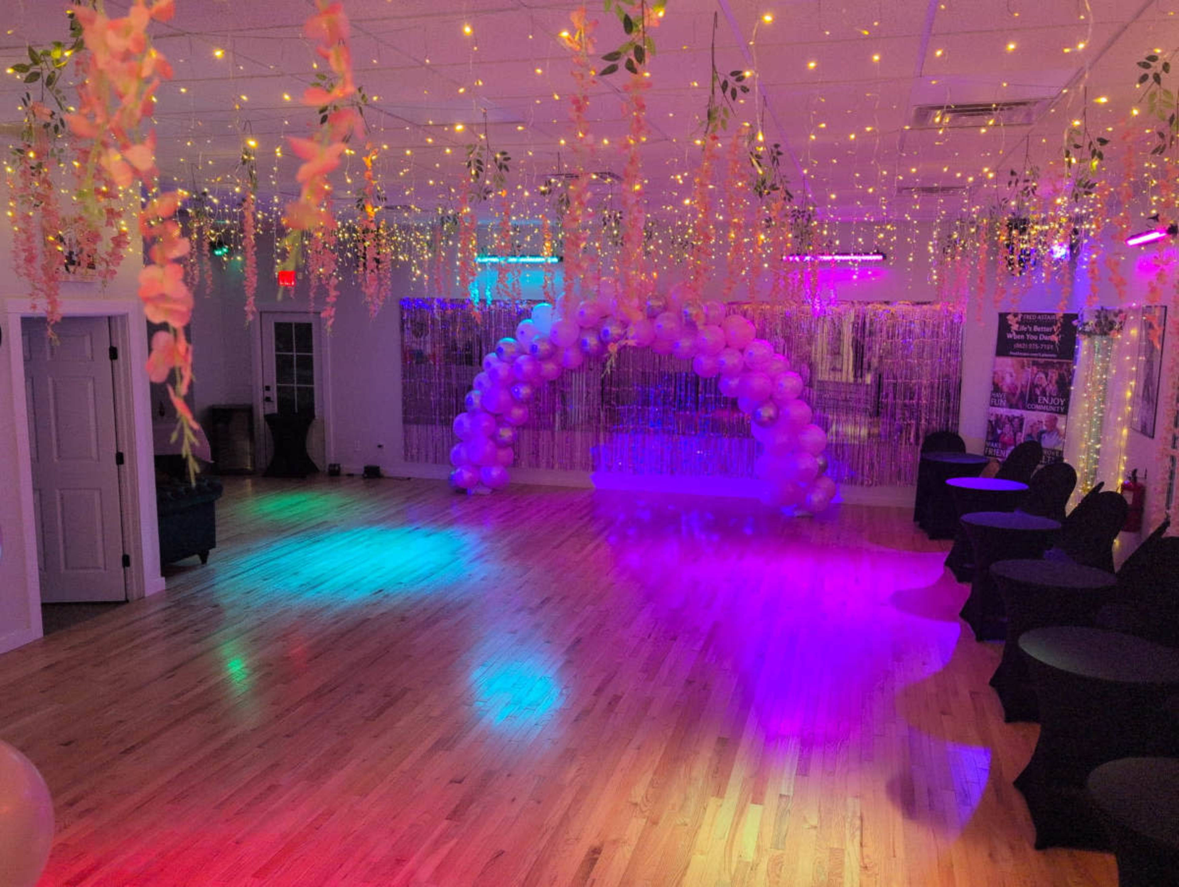 The image shows a decorated event space with colorful lights, an arch made of balloons, and tables draped in black cloth.