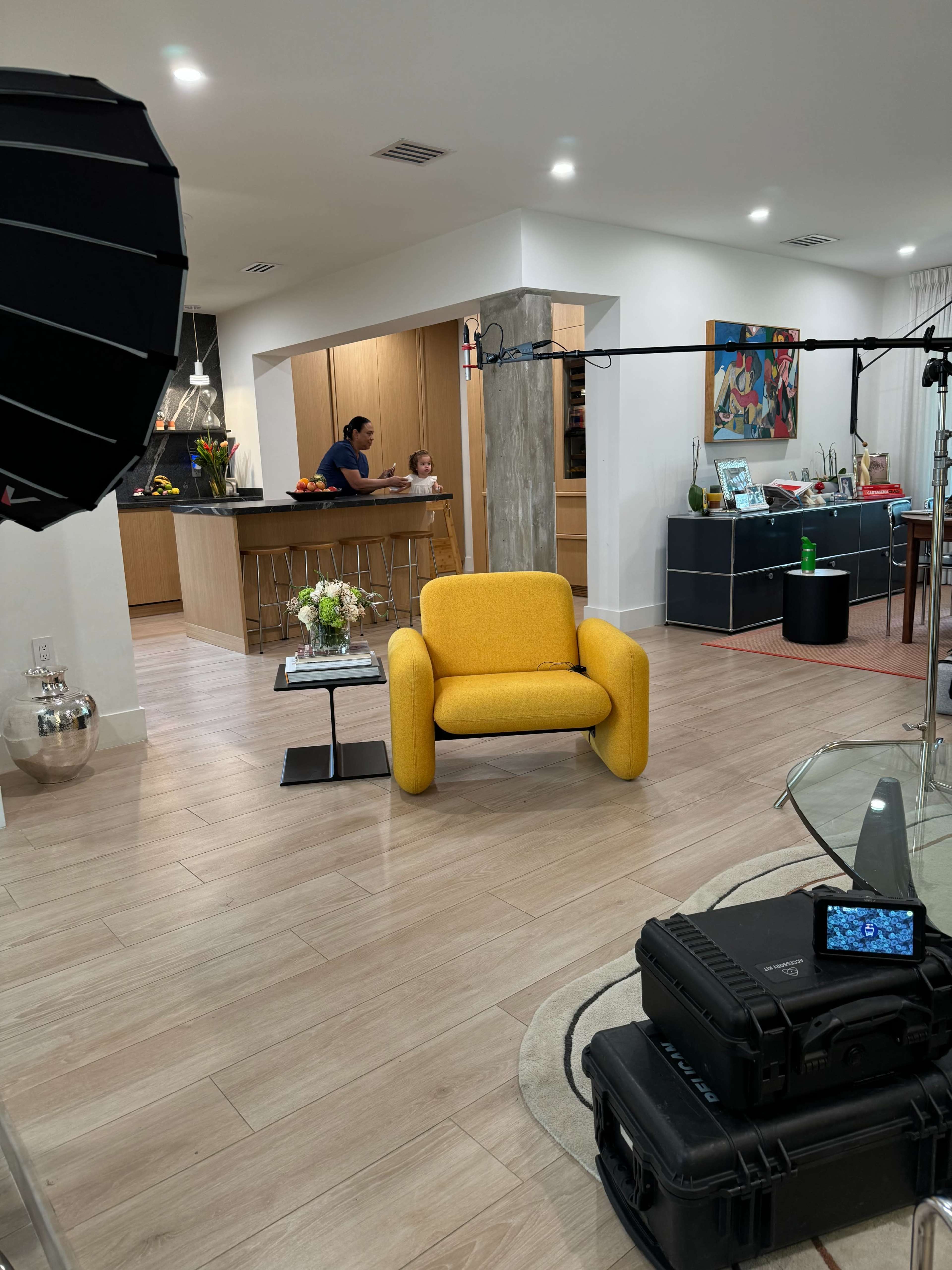 A yellow armchair sits in the foreground of a modern, open-concept kitchen and living area with filming equipment set up nearby.